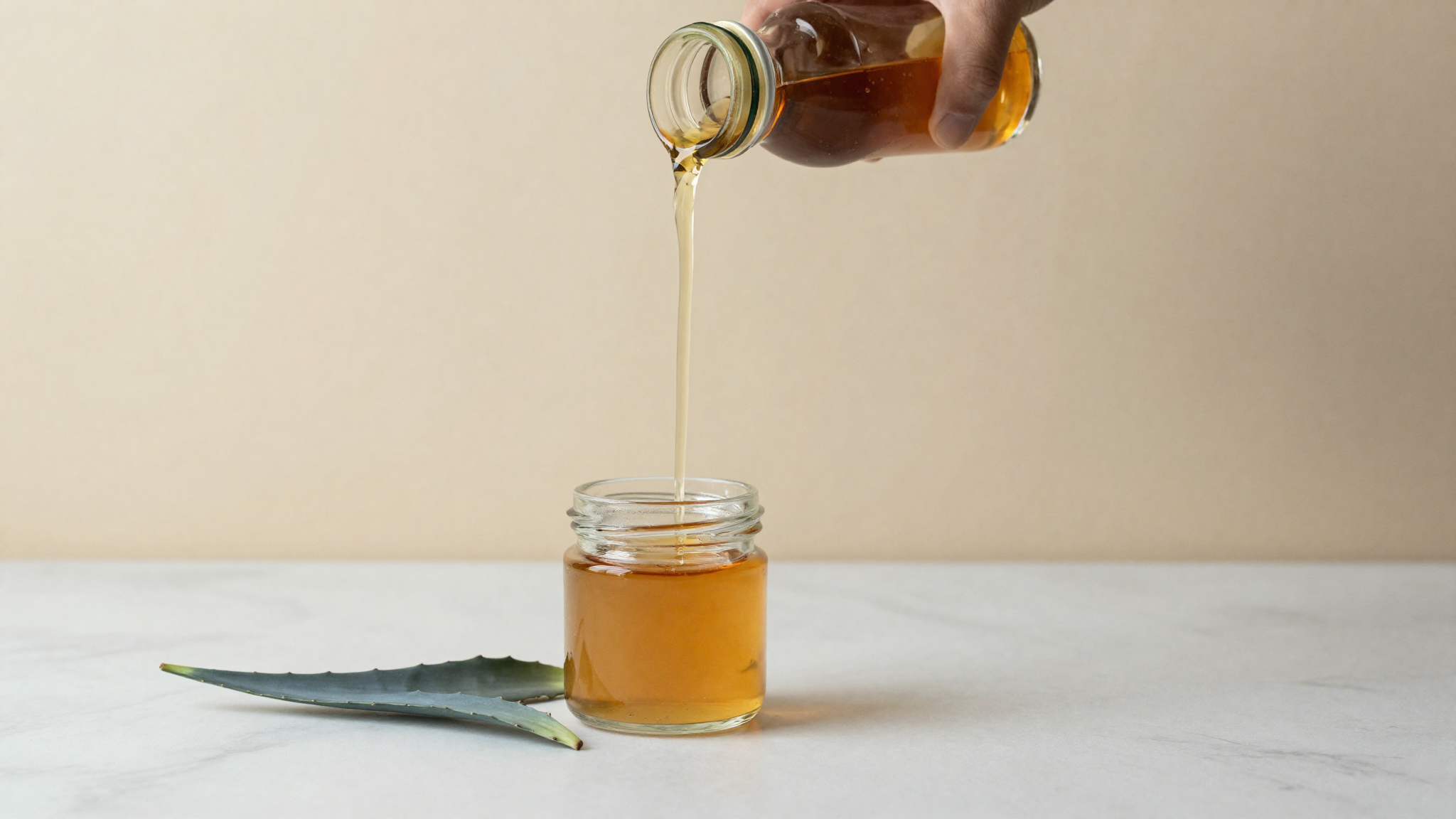 Amber agave syrup pouring into a glass jar beside a blue agave leaf on marble