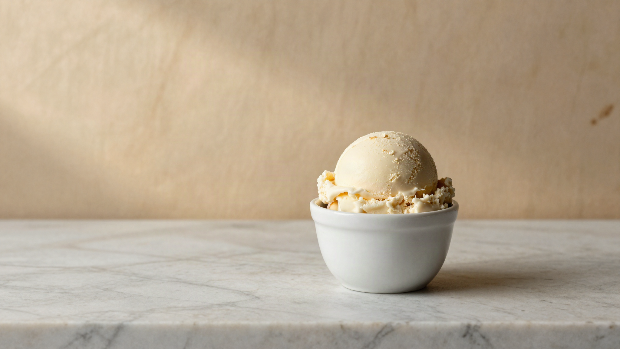 A scoop of pale ivory almond gelato in a white ceramic cup on marble