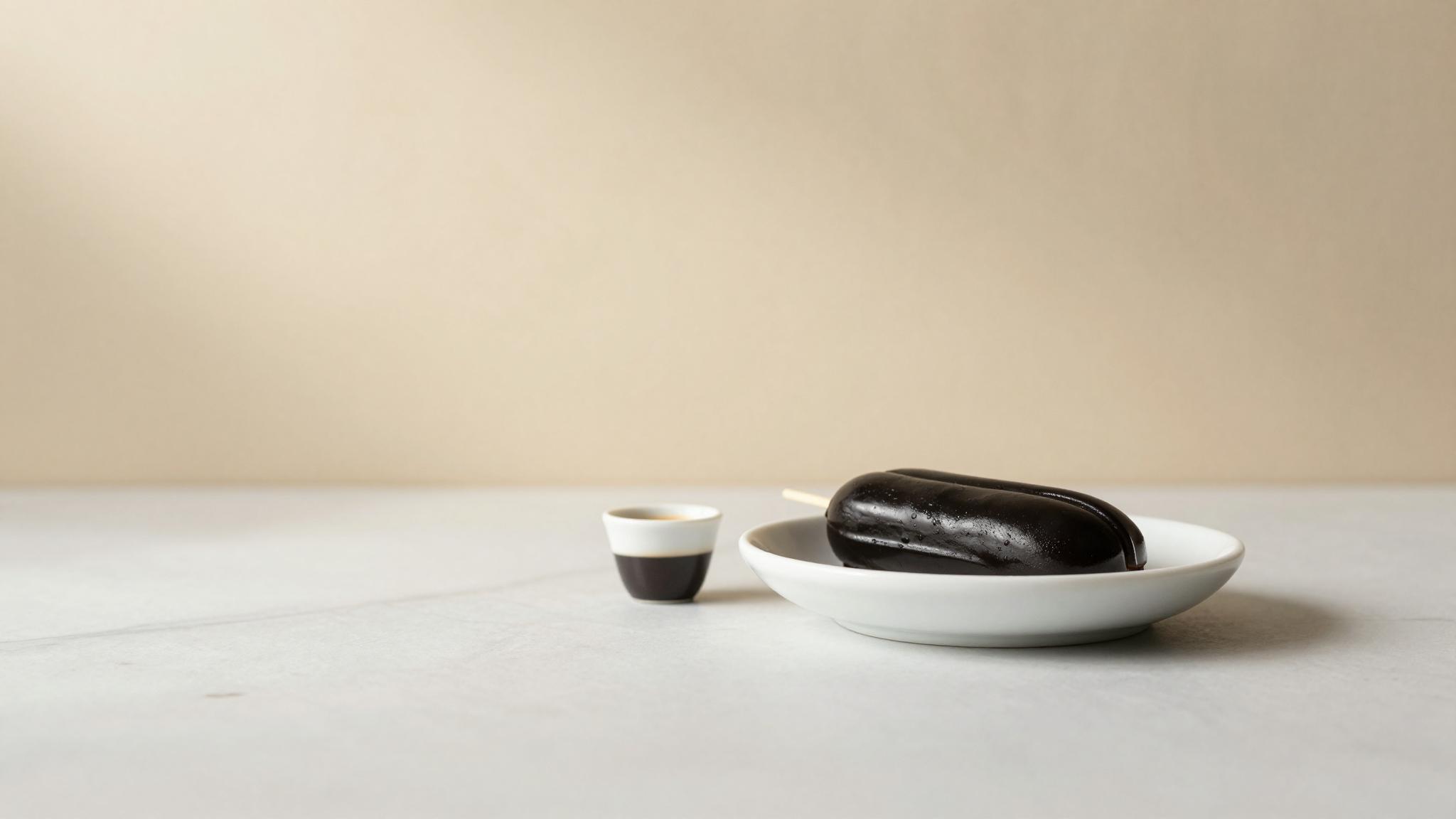 Closing still life of a glossy black licorice gelato quenelle in a white ceramic dish with a small espresso cup nearby