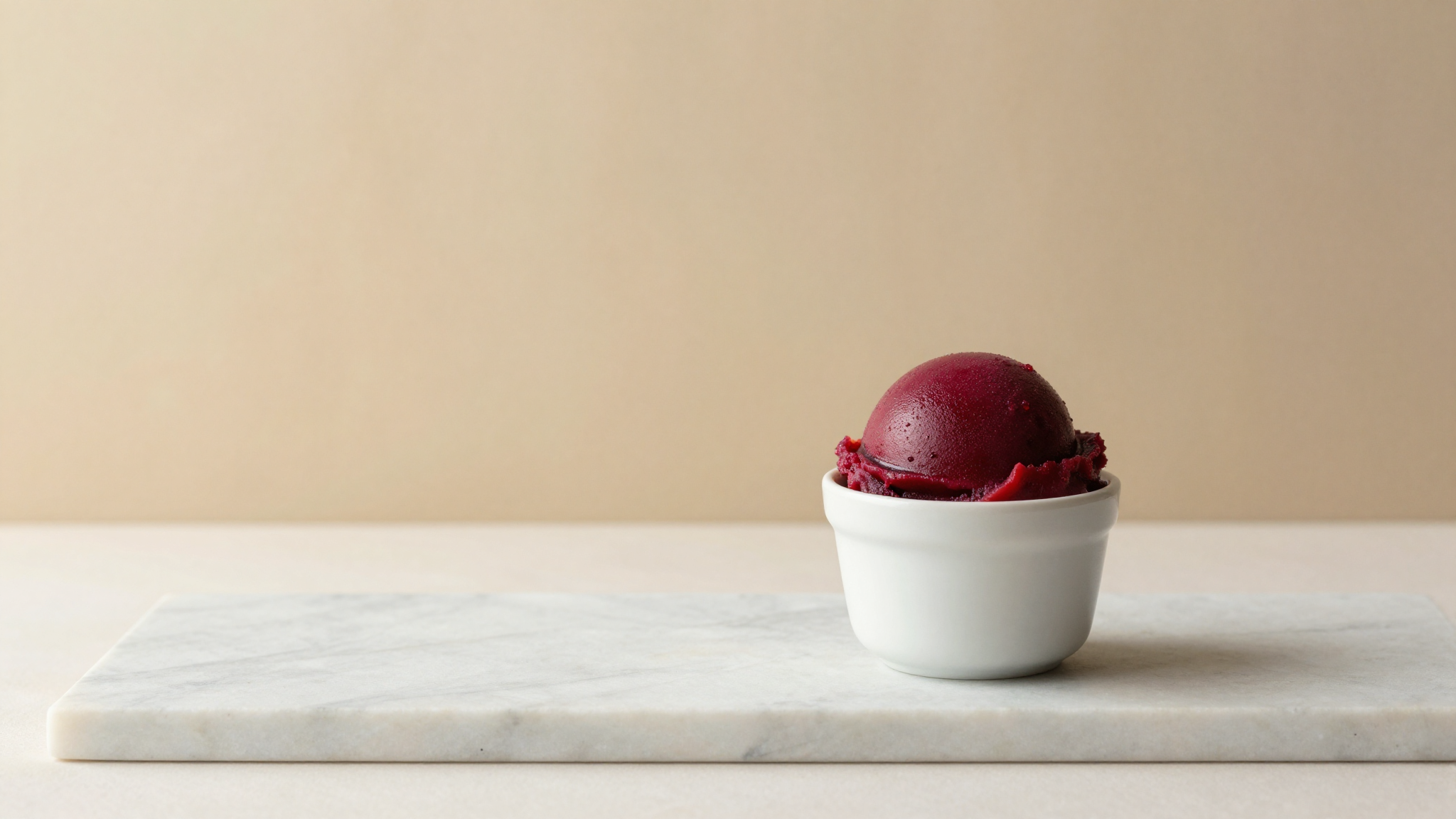 A scoop of deep red blood orange sorbet in a white ceramic cup on a marble counter