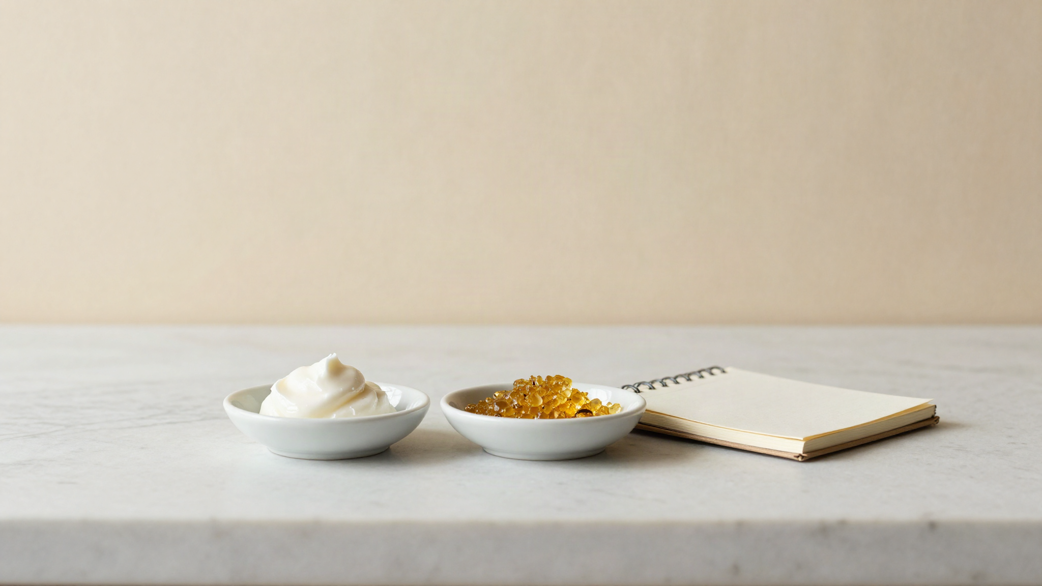 Two small white ceramic dishes on marble, one with waxy white emulsifier flakes and one with golden lecithin granules, beside a small open notebook