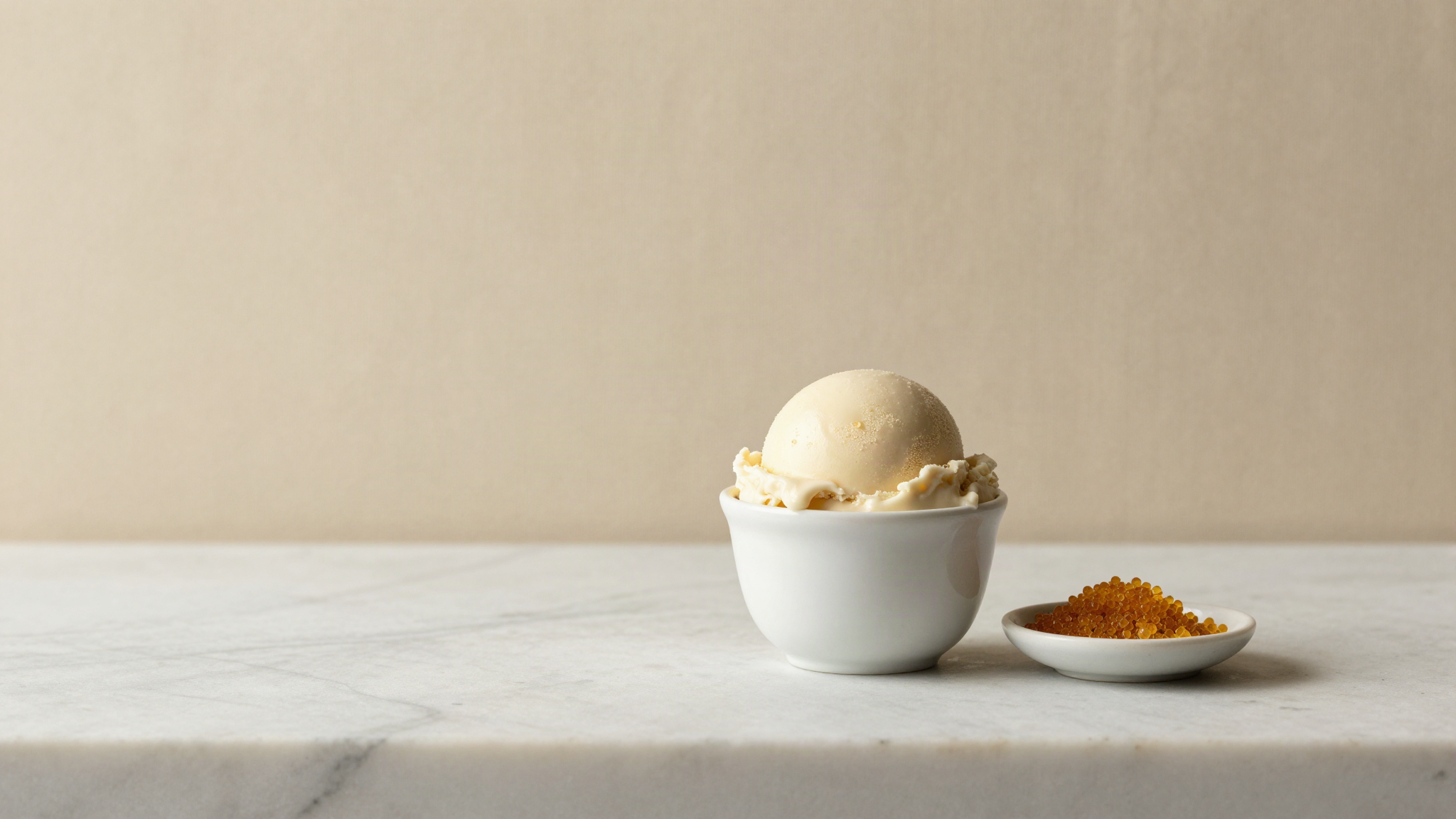 A single smooth scoop of pale vegan gelato in a white ceramic cup on a marble counter beside a small dish of golden lecithin granules
