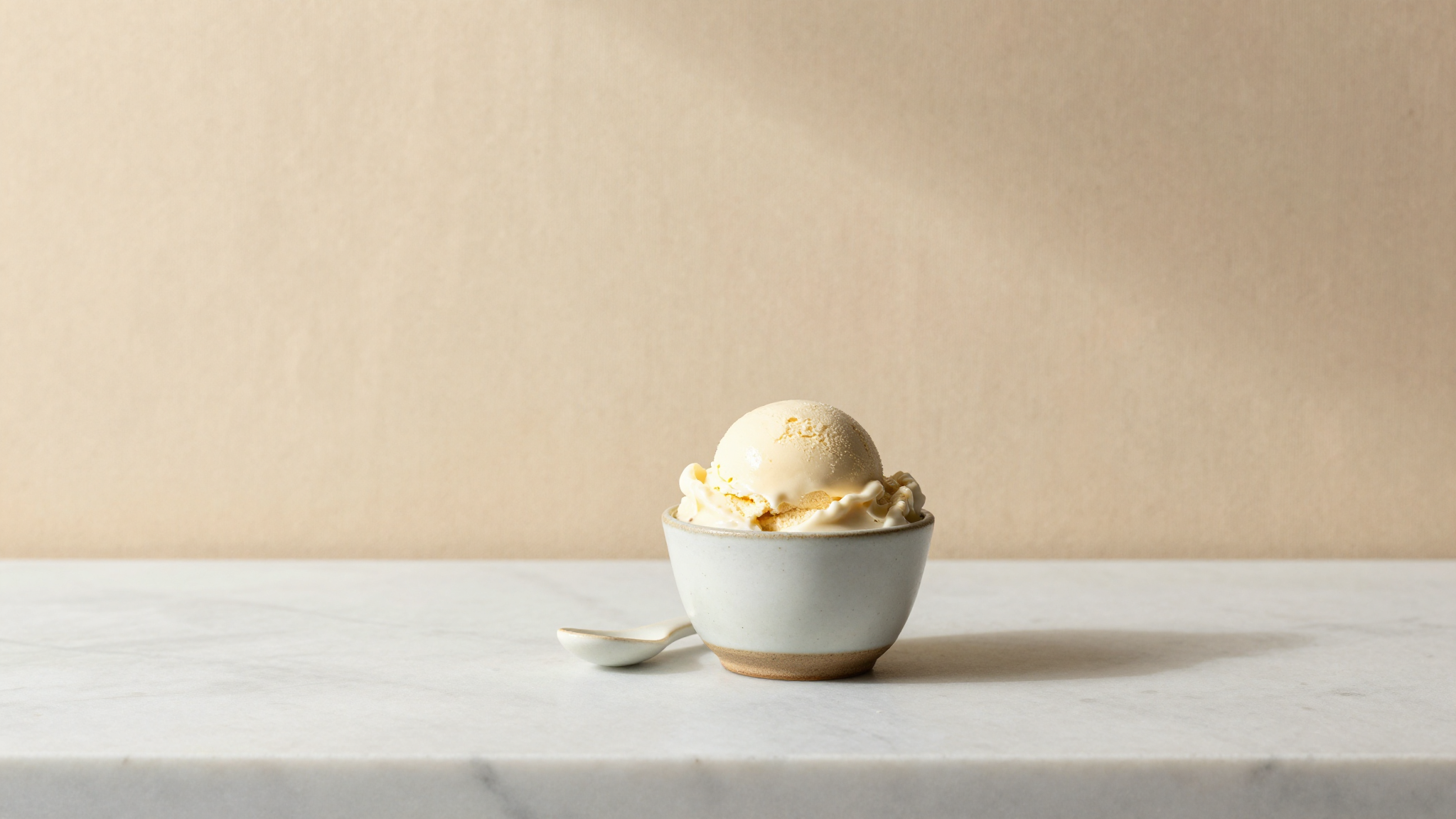 A small ceramic cup of fresh creamy homemade gelato with a tiny ceramic spoon beside it on a clean white marble counter