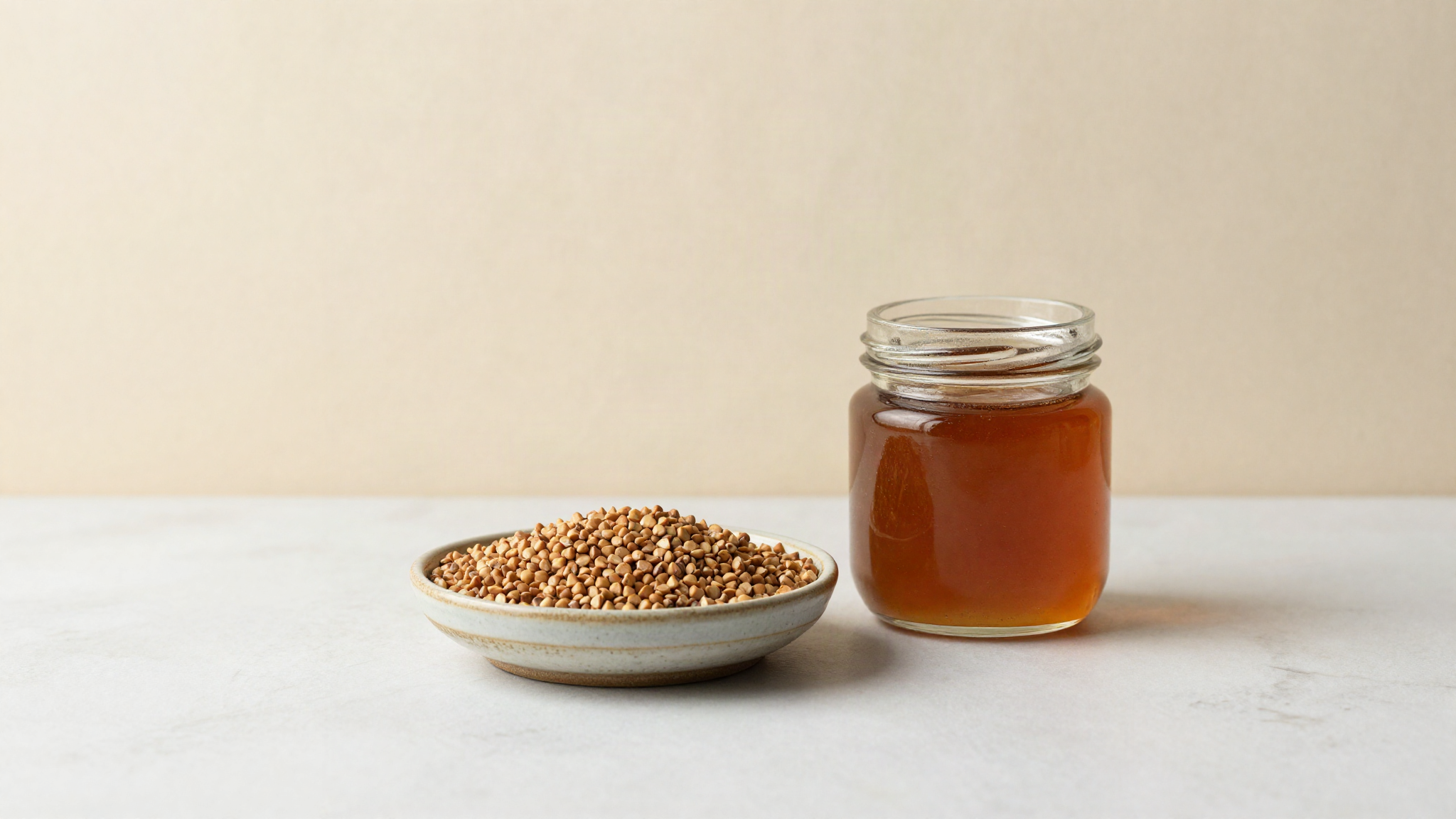 Raw buckwheat groats in a ceramic dish beside a jar of dark amber honey on white marble