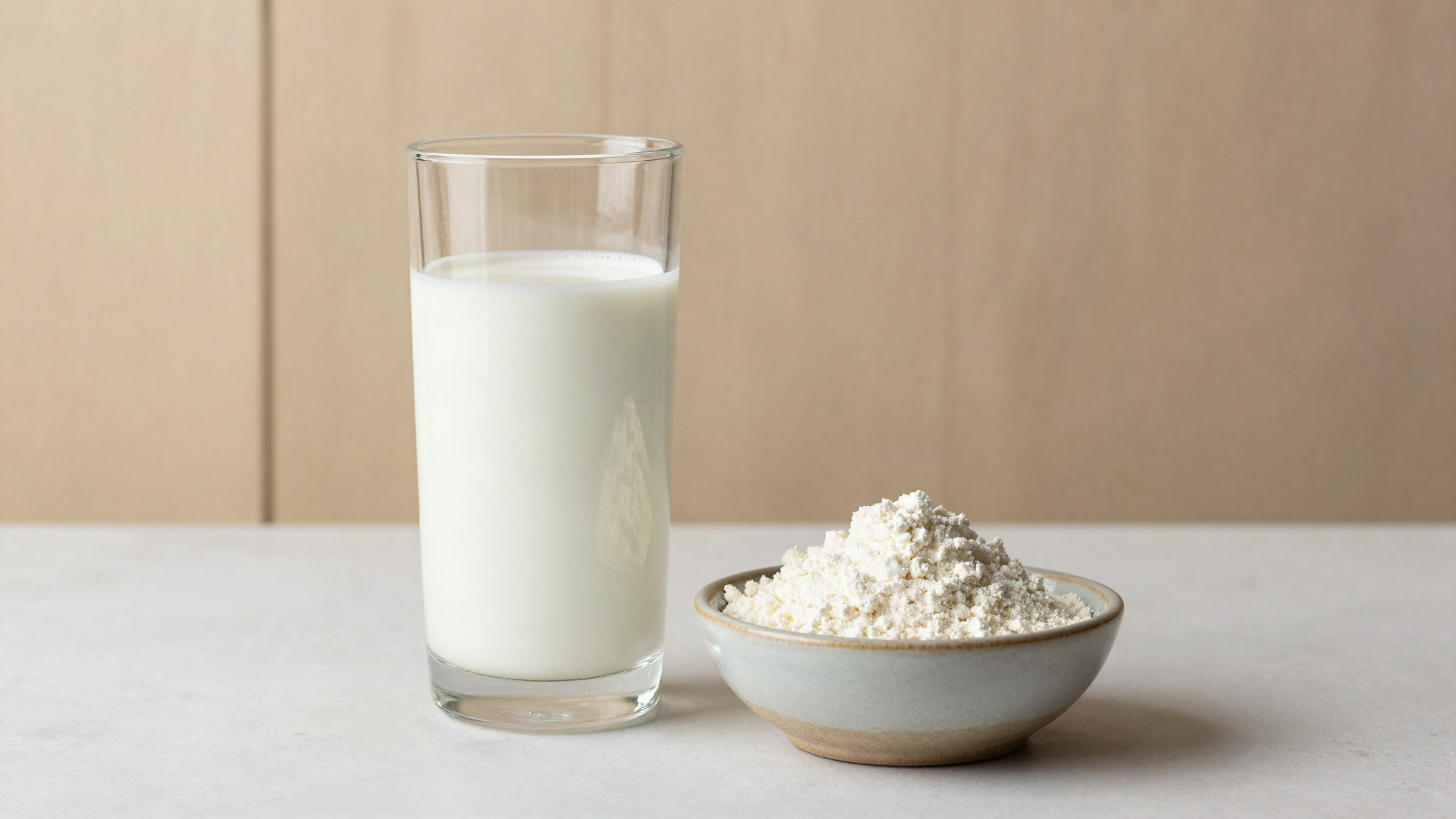 A tall glass of cold whole milk beside a small ceramic bowl of fine white milk protein powder on a pale counter