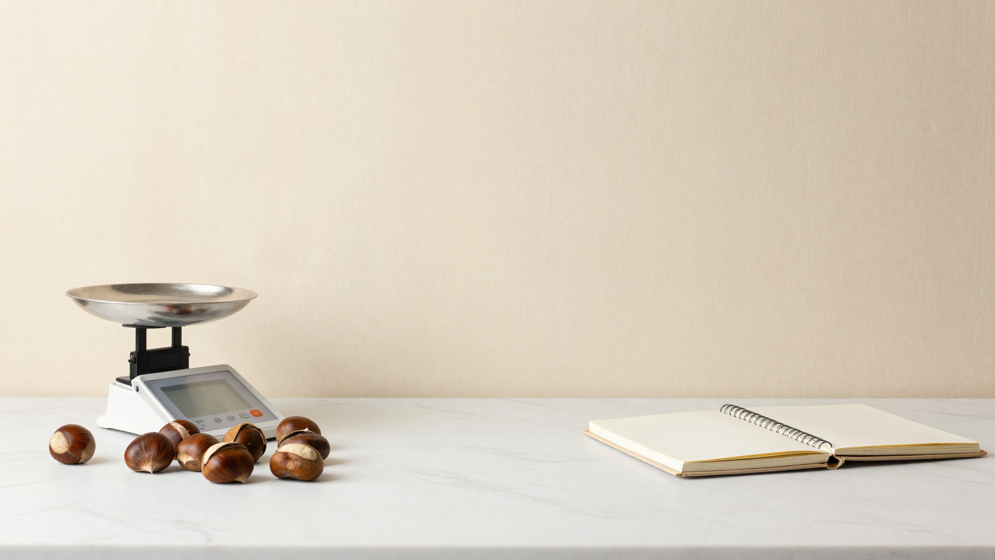 Whole raw chestnuts beside a small kitchen scale and a notebook on marble