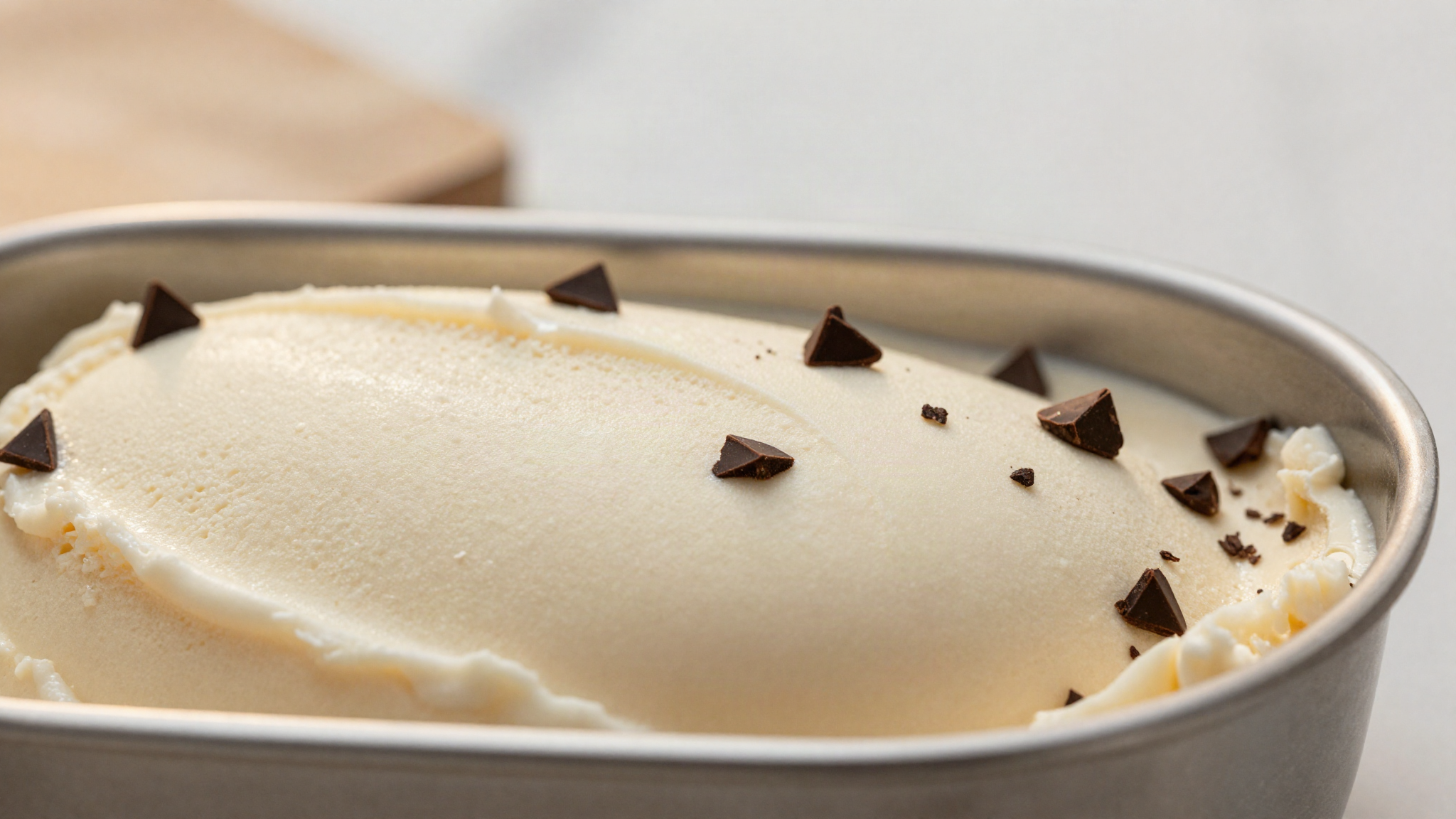 Macro close-up of dark chocolate chips embedded in the smooth pale surface of gelato in a steel pan