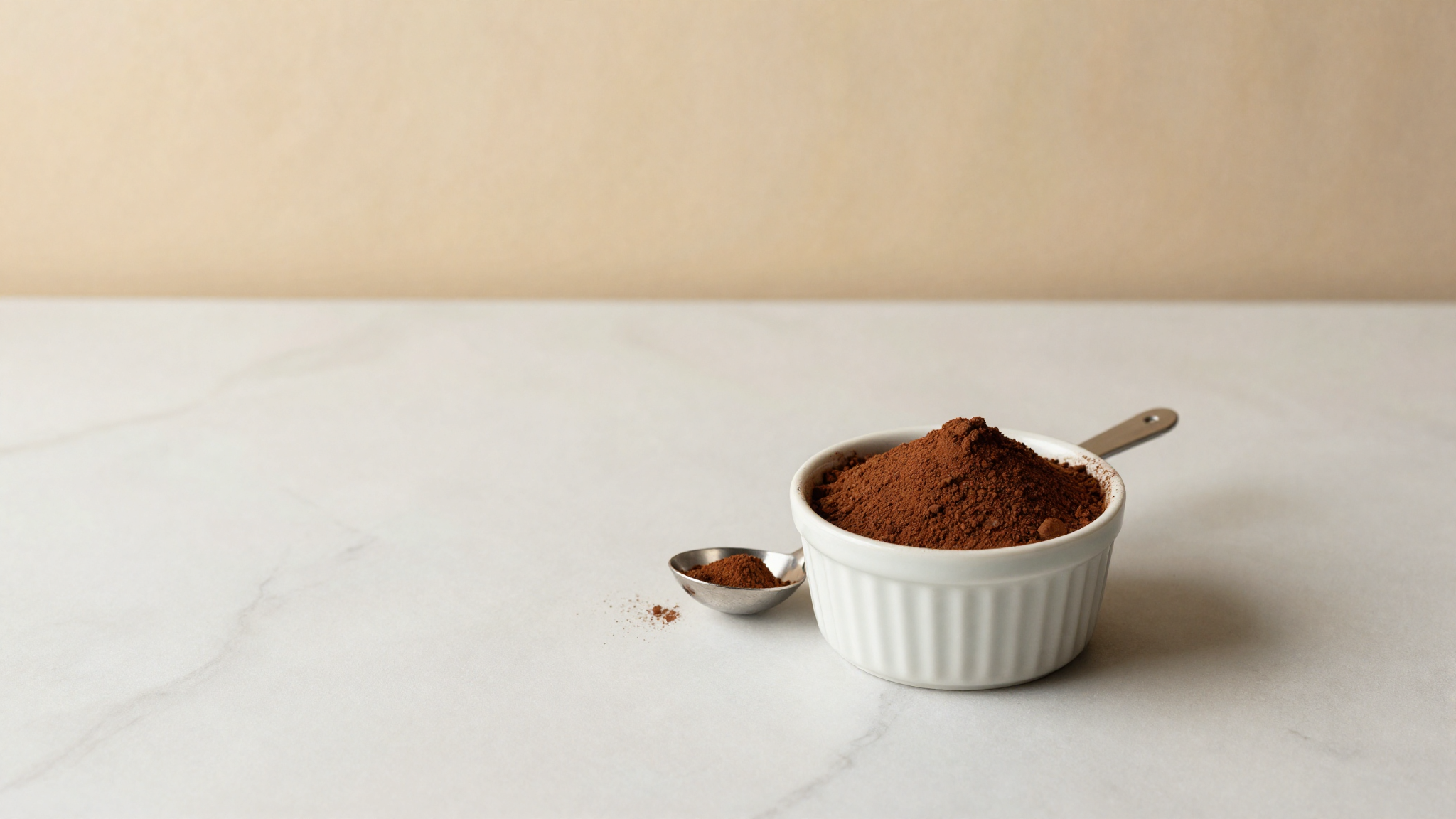 Top-down porcelain ramekin filled with rich dark cocoa powder beside a small silver scoop on marble
