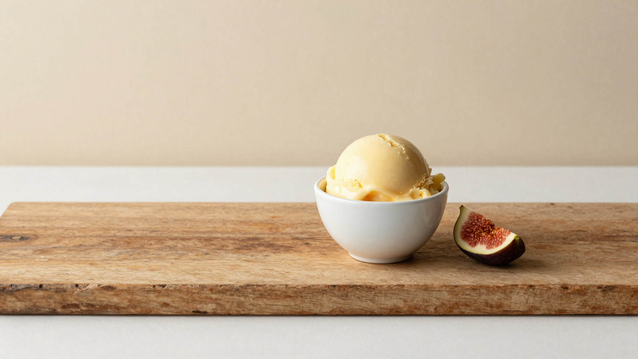 Finished crema all'uovo gelato — pale yellow scoop in white ceramic cup on aged wood with a fresh fig leaf, italian artisan presentation