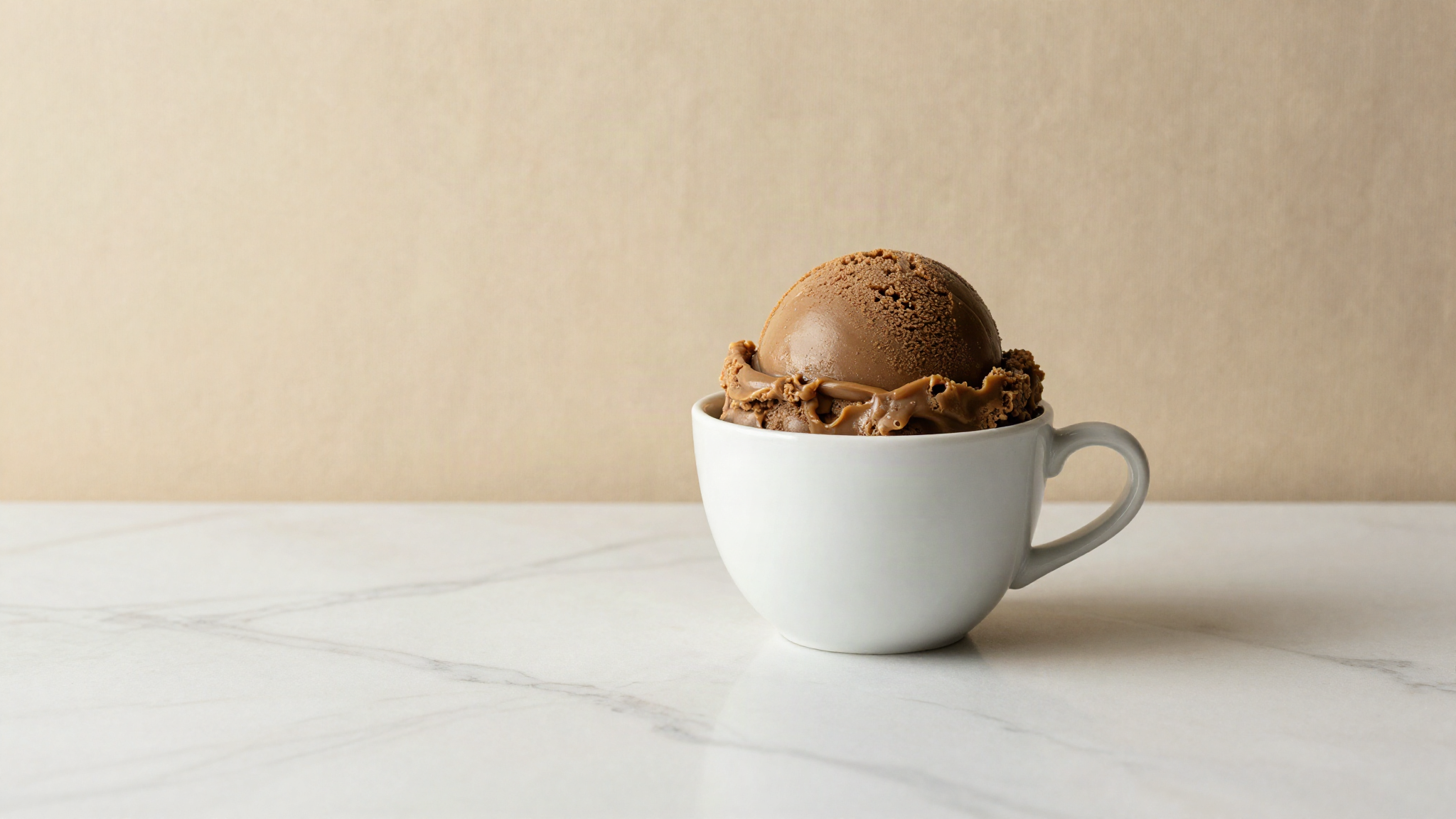 A scoop of dark caramel toned gelato in a white ceramic cup on marble