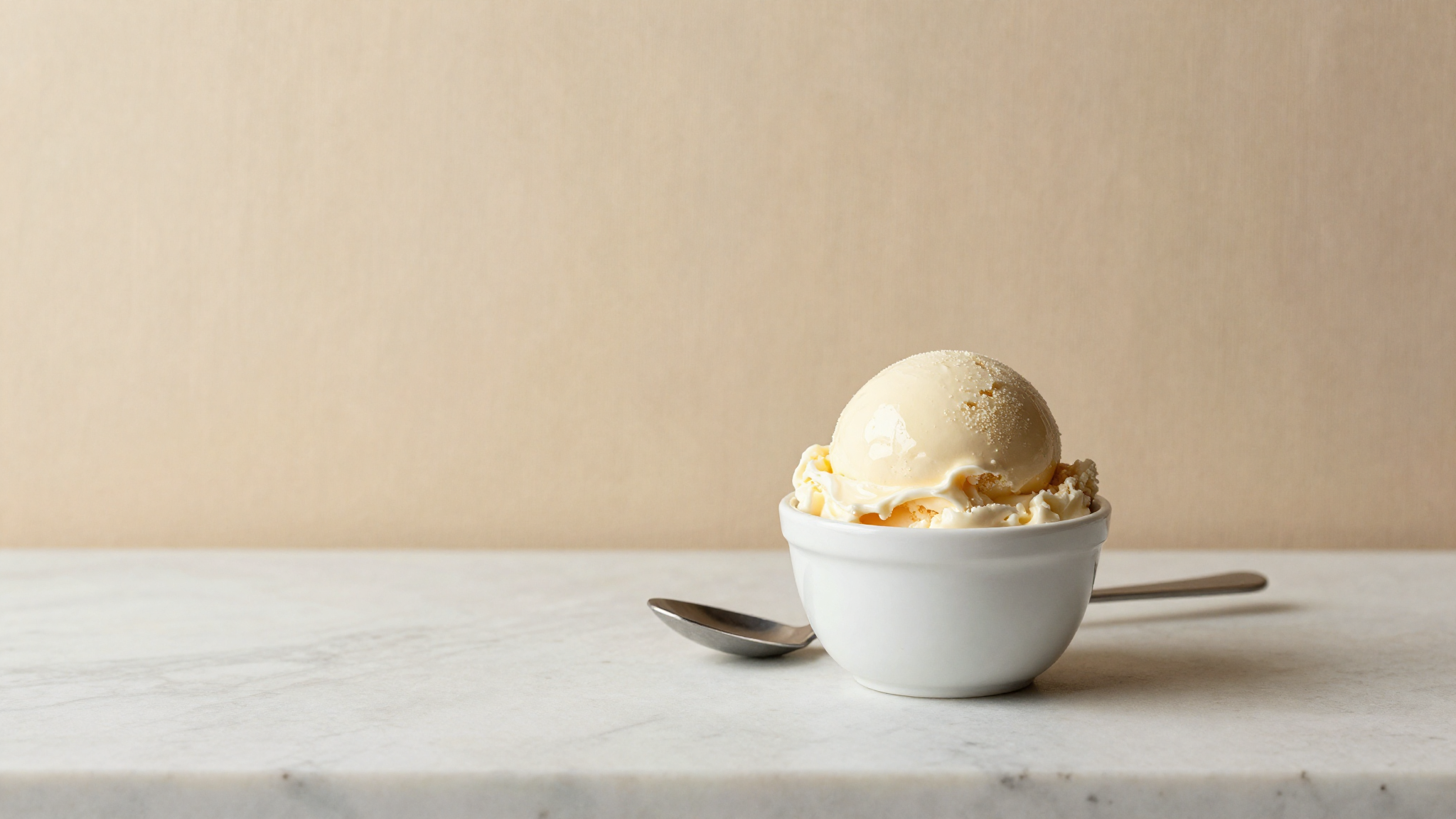 A single glossy scoop of pale cream gelato in a white ceramic cup with a metal spade beside it