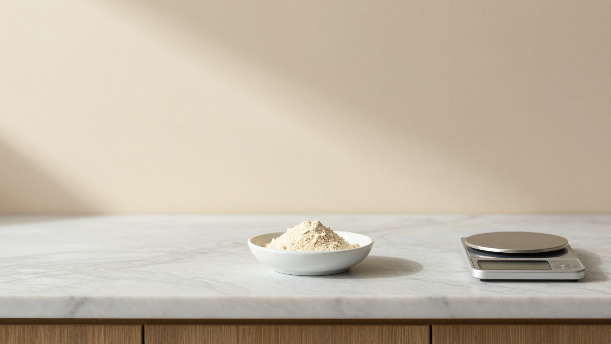 A small white ceramic dish containing pale beige stabilizer powder beside a tiny digital scale and a notebook on a marble counter