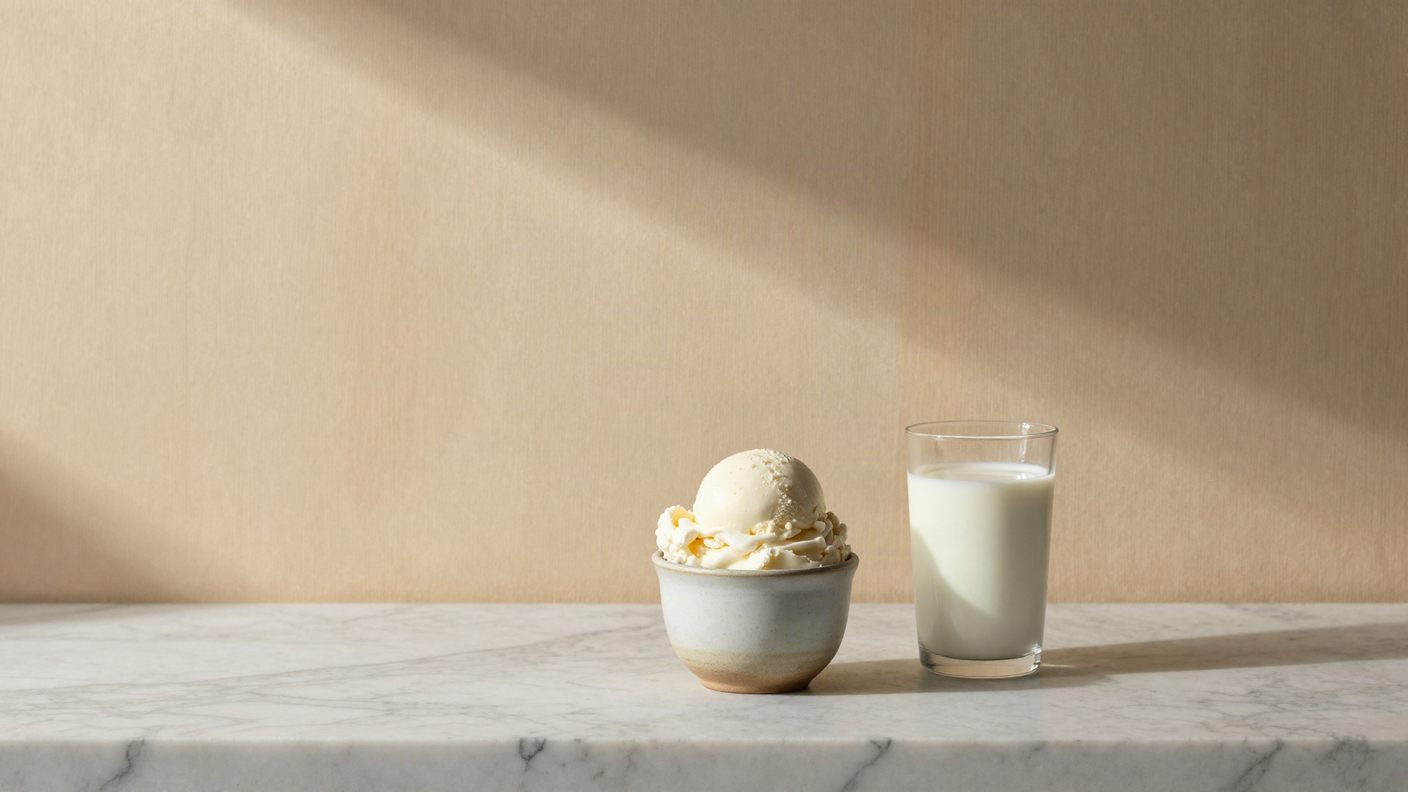 A single scoop of white fior di latte gelato in a small ceramic cup on a marble counter beside a glass of milk