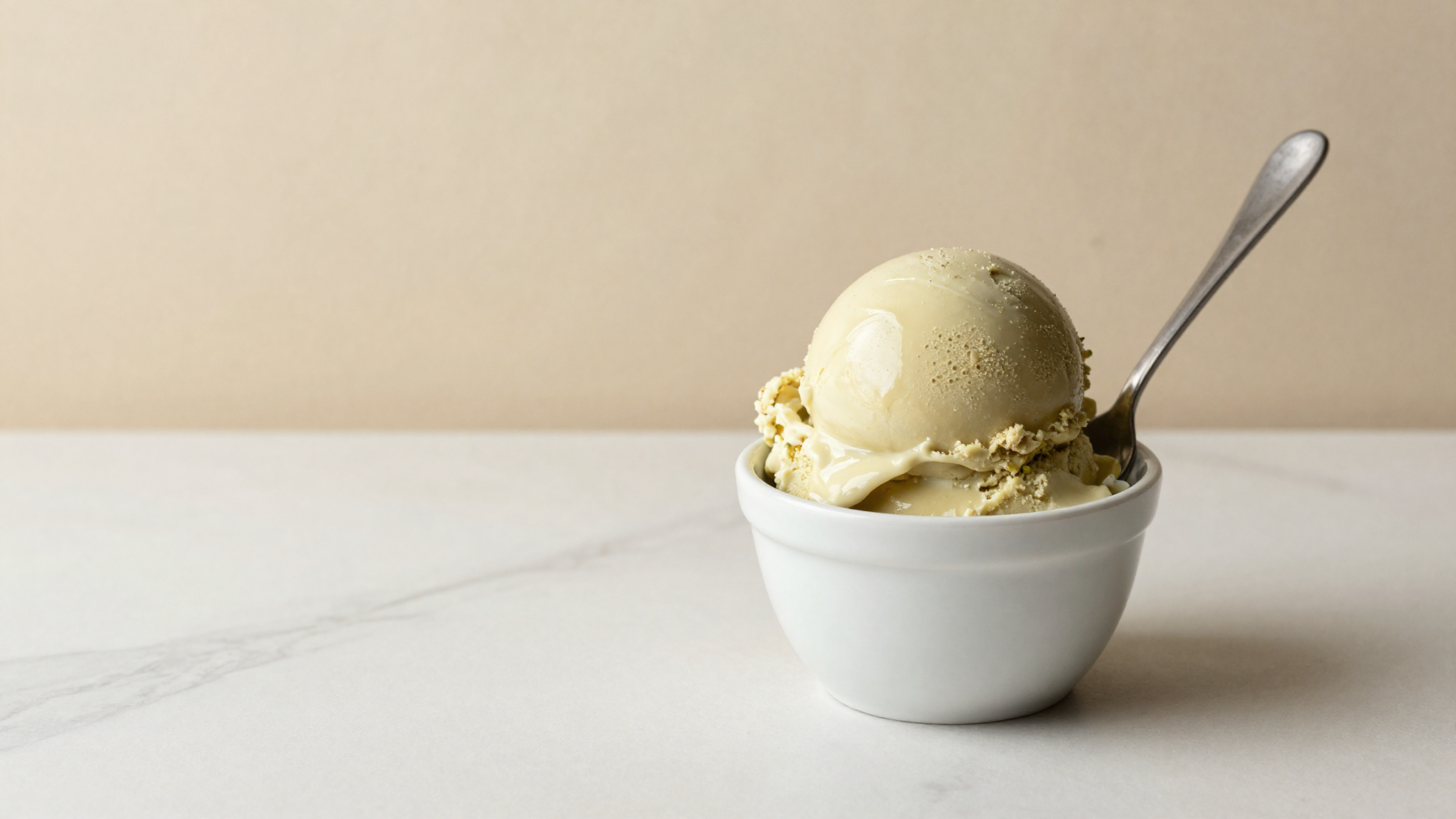 A single glossy scoop of pale gelato in a white ceramic cup with a metal spade beside it