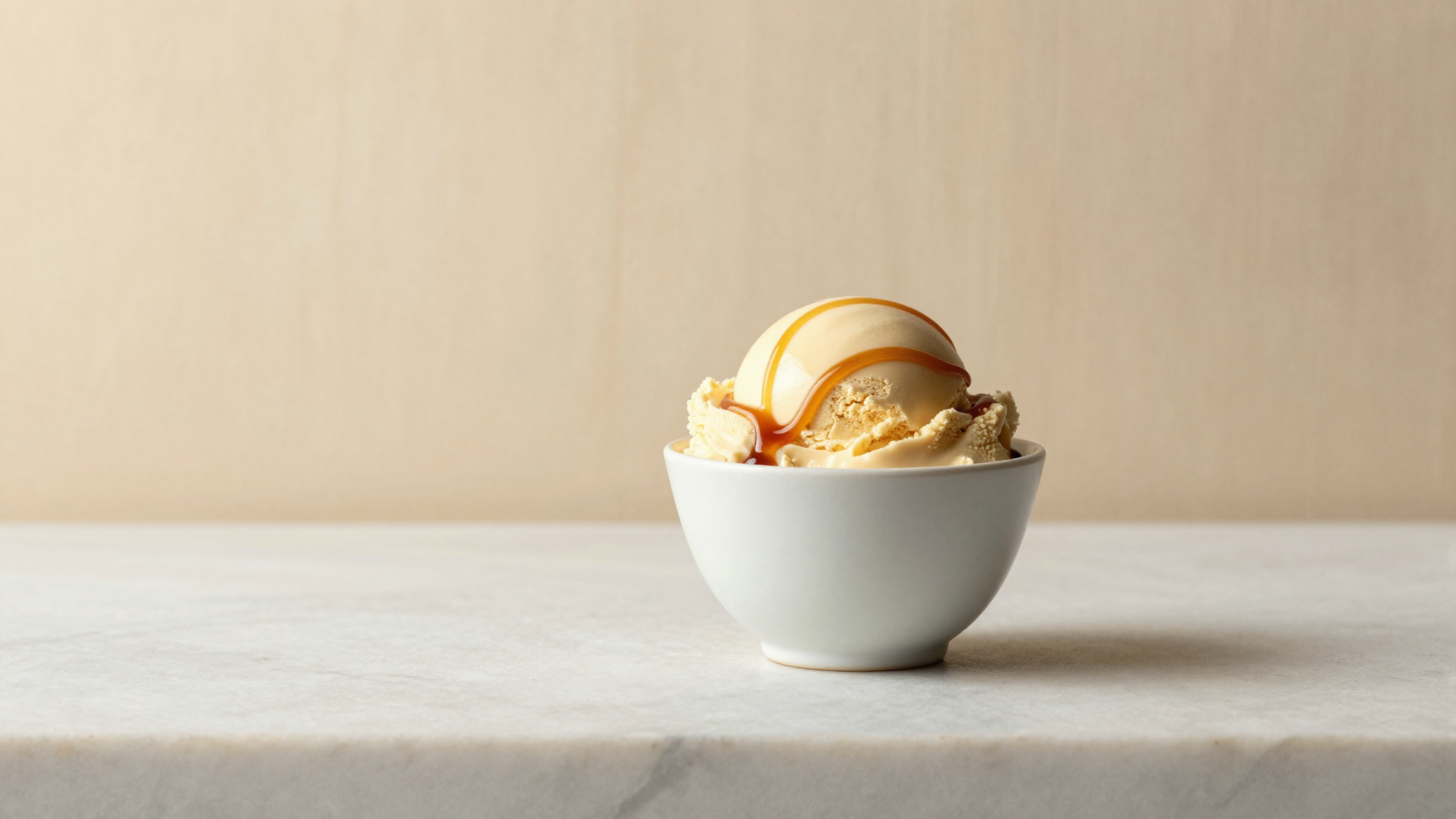 A serving of caramel coloured dulce de leche gelato in a white ceramic cup on marble