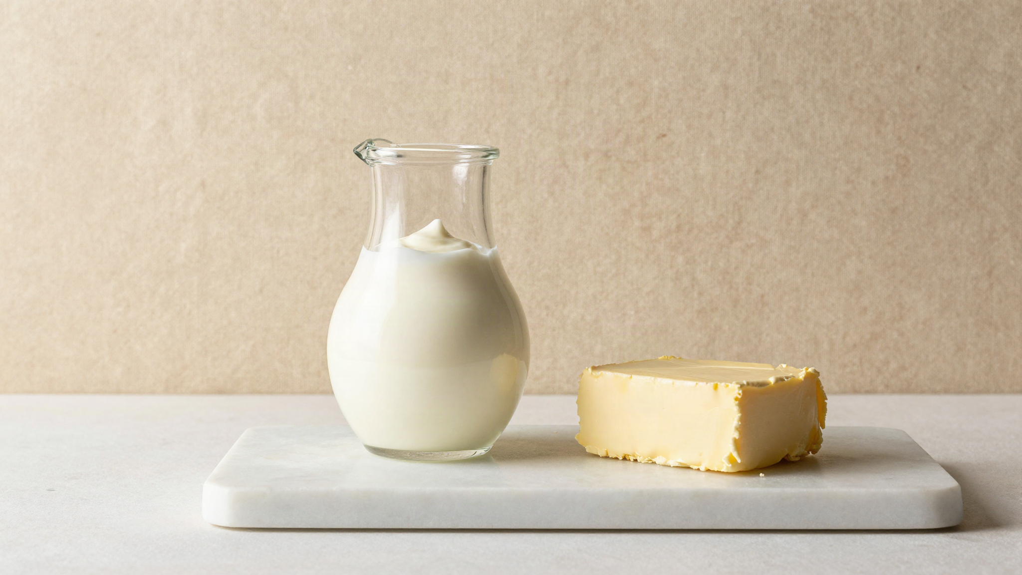 A glass jug of fresh cream beside a block of pale butter on white marble