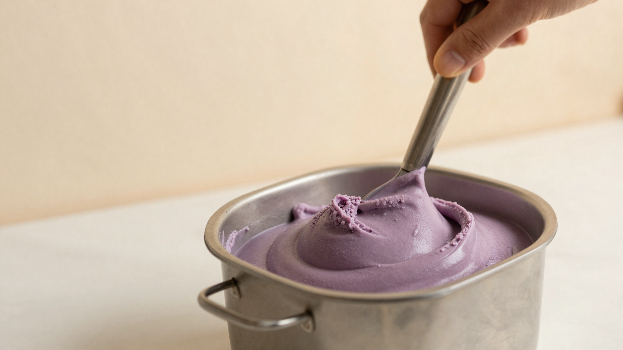 Fig gelato mix churning in a curved stainless pan with a single paddle developing a soft violet hue