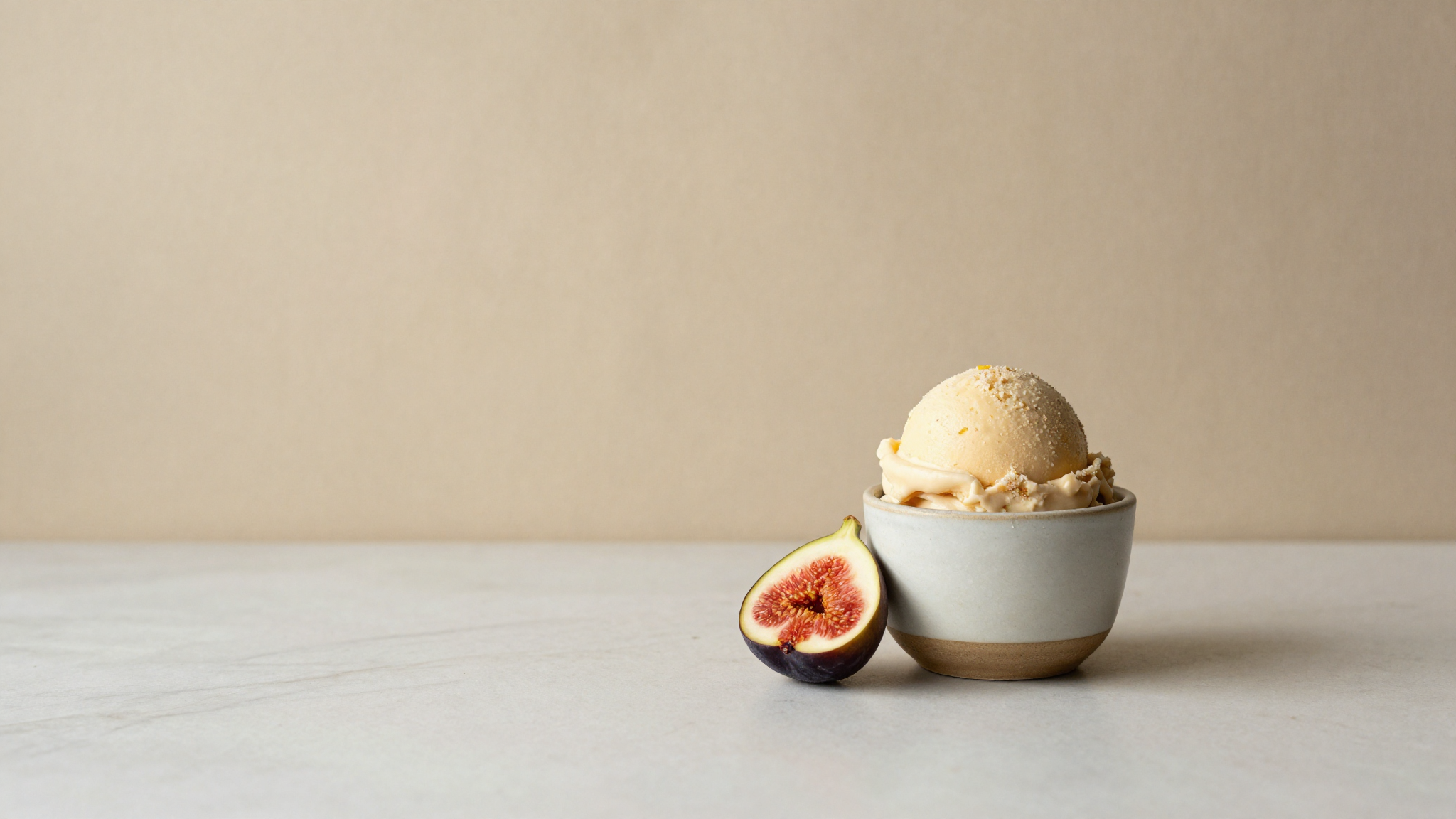 Single scoop of fig gelato in a ceramic cup with one fresh fig half beside it on marble
