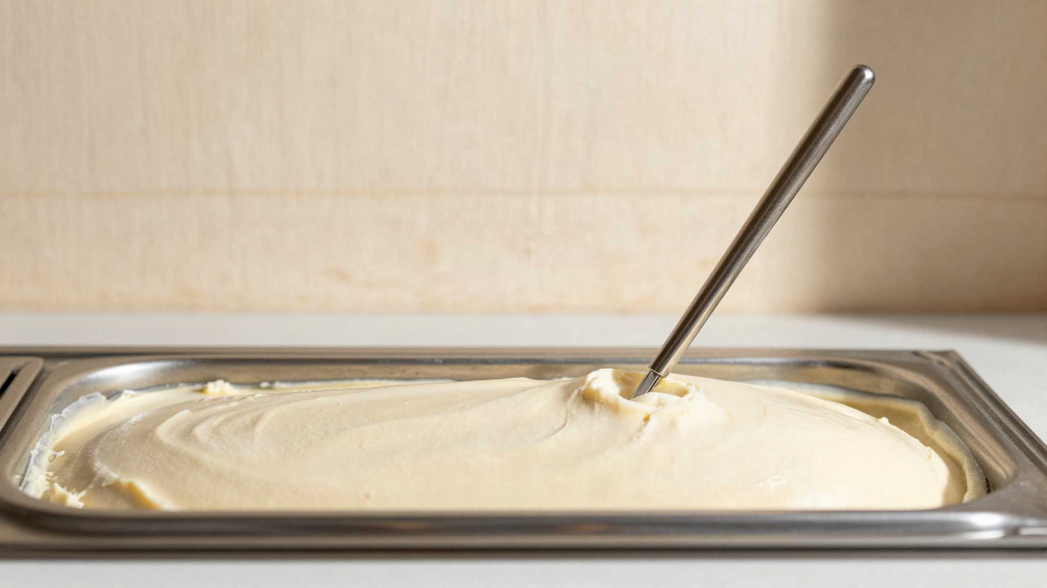 Stainless steel probe thermometer inserted into the surface of pale gelato in a display case pan