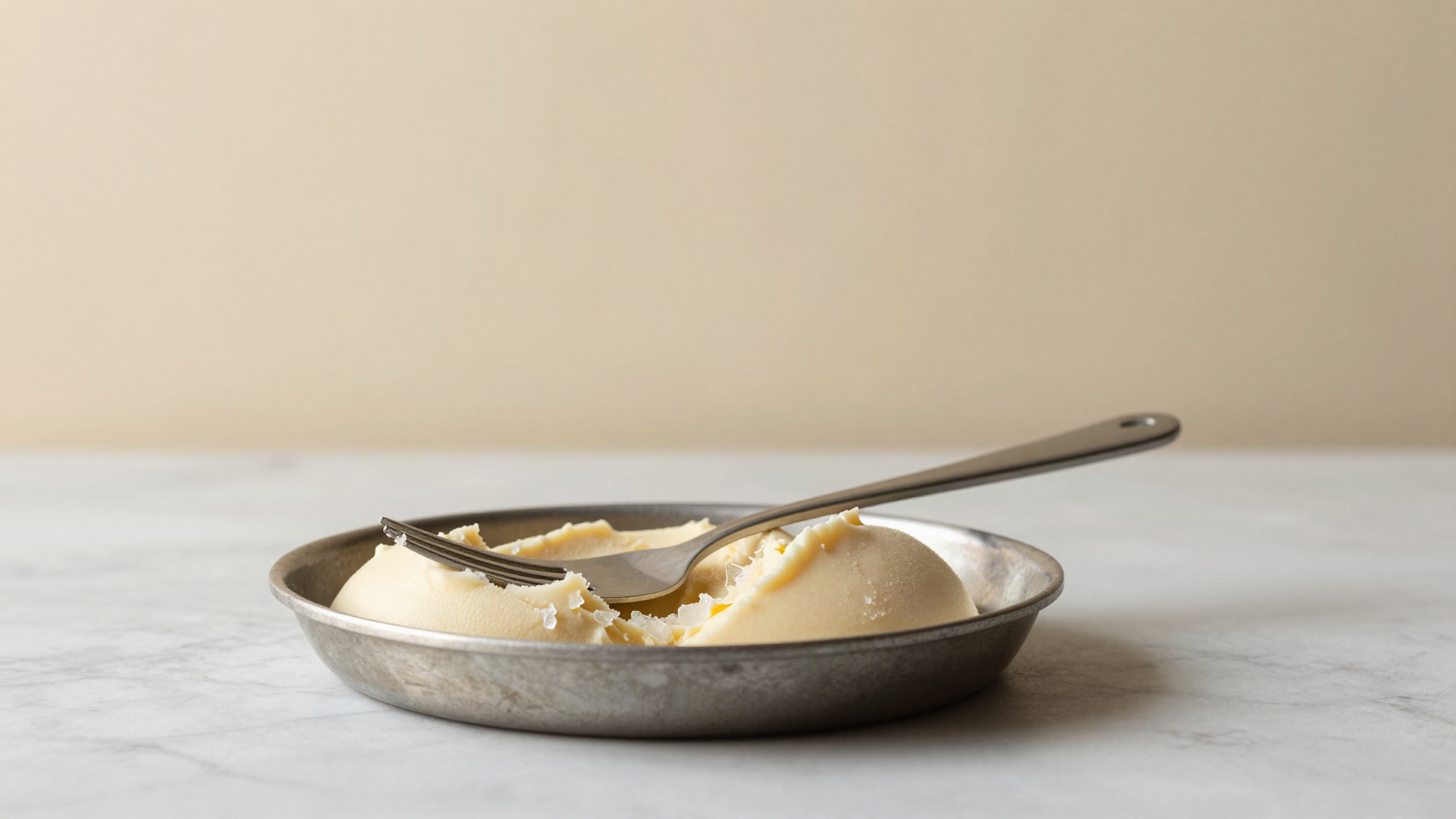 A wide shallow metal pan of pale gelato base with frosted edges beside a fork and a stiff spatula on marble