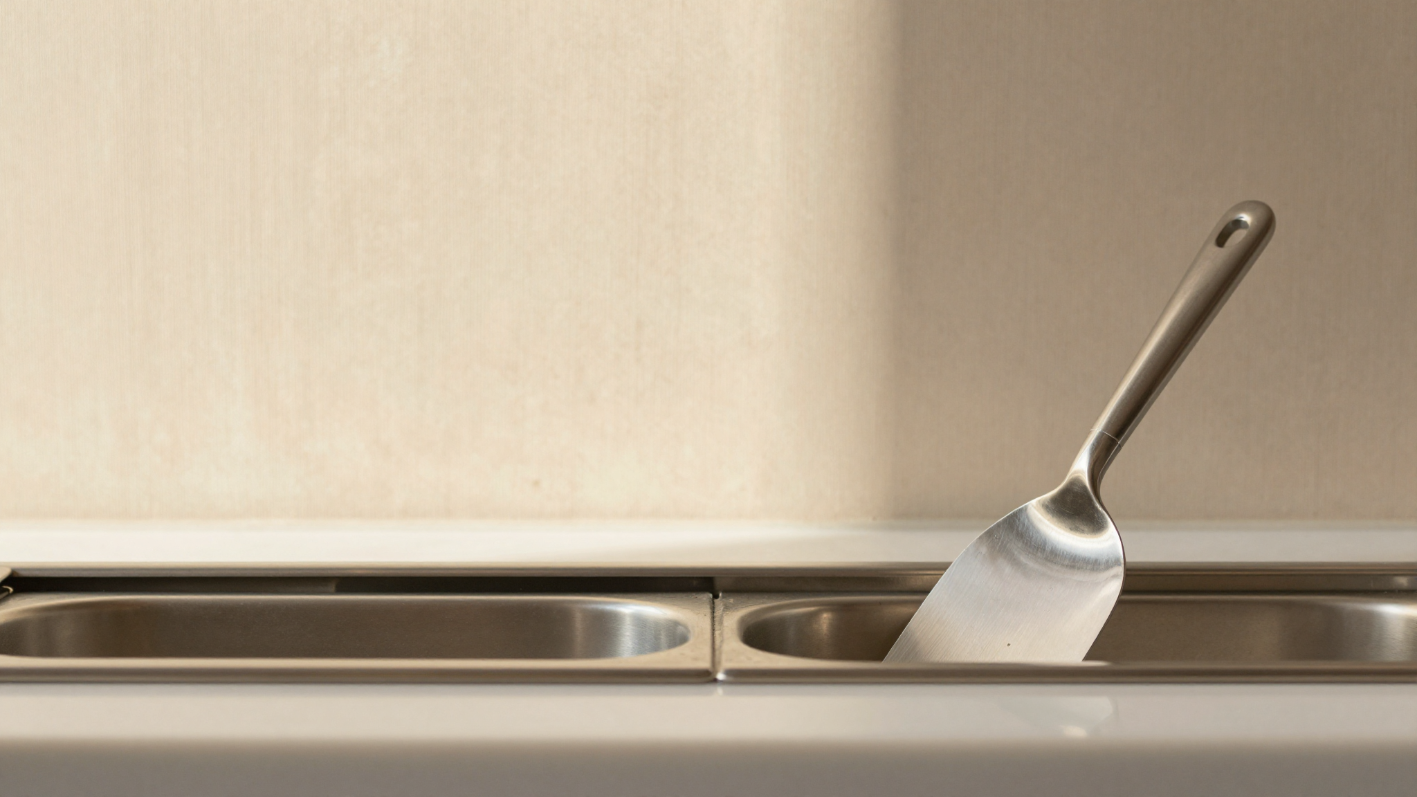 Close-up of polished stainless steel gelato spatola resting against the rim of a stainless steel gelato pan inside a vetrina
