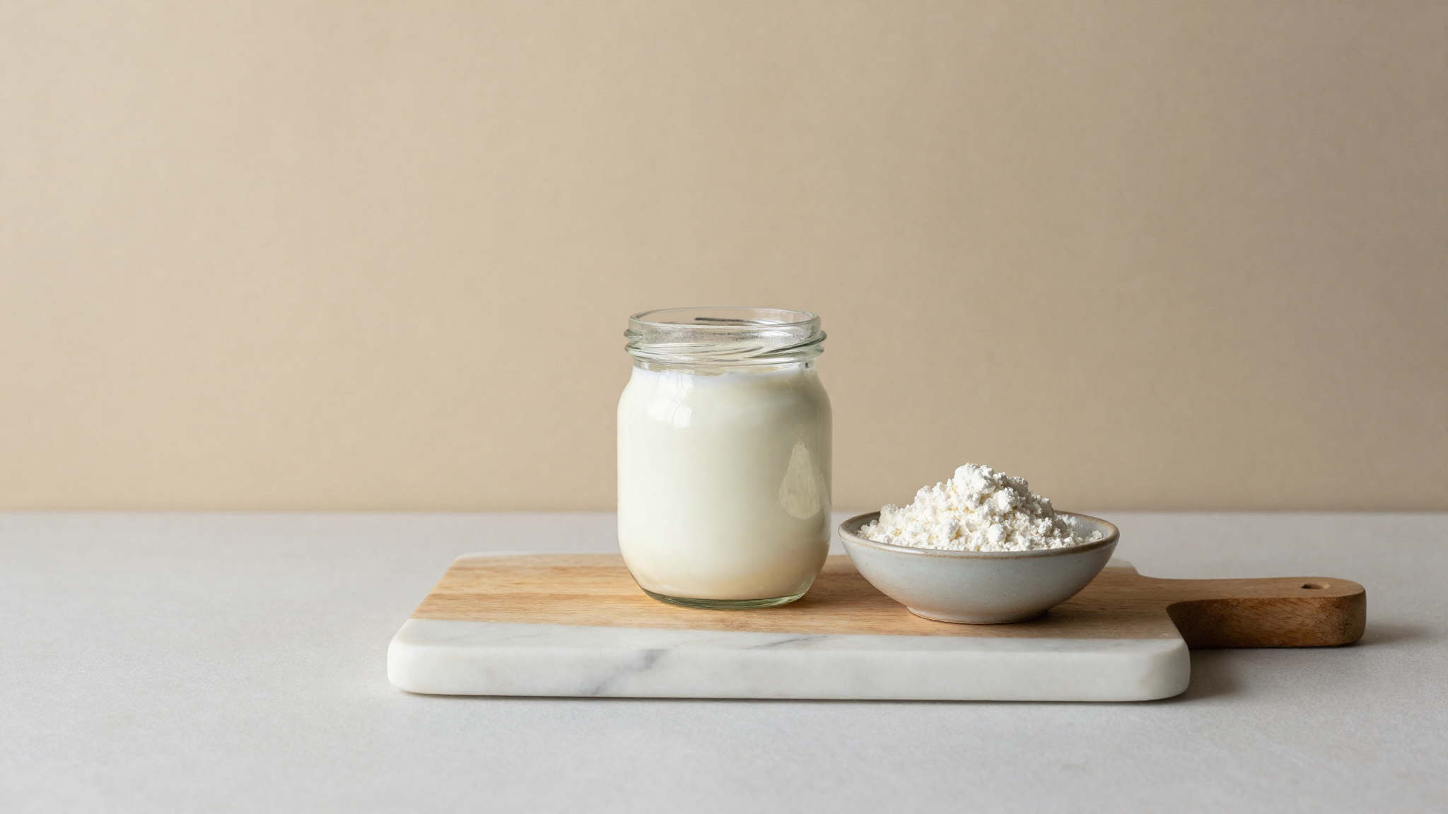 A wooden serving board with a glass jar of plain yogurt and a ceramic dish of milk powder on white marble
