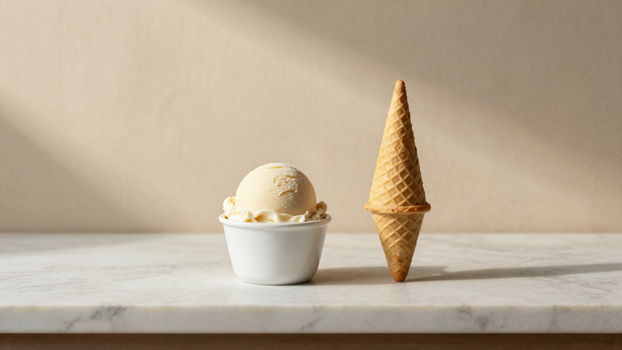 A scoop of pale gelato in a white ceramic cup beside a waffle cone on white marble