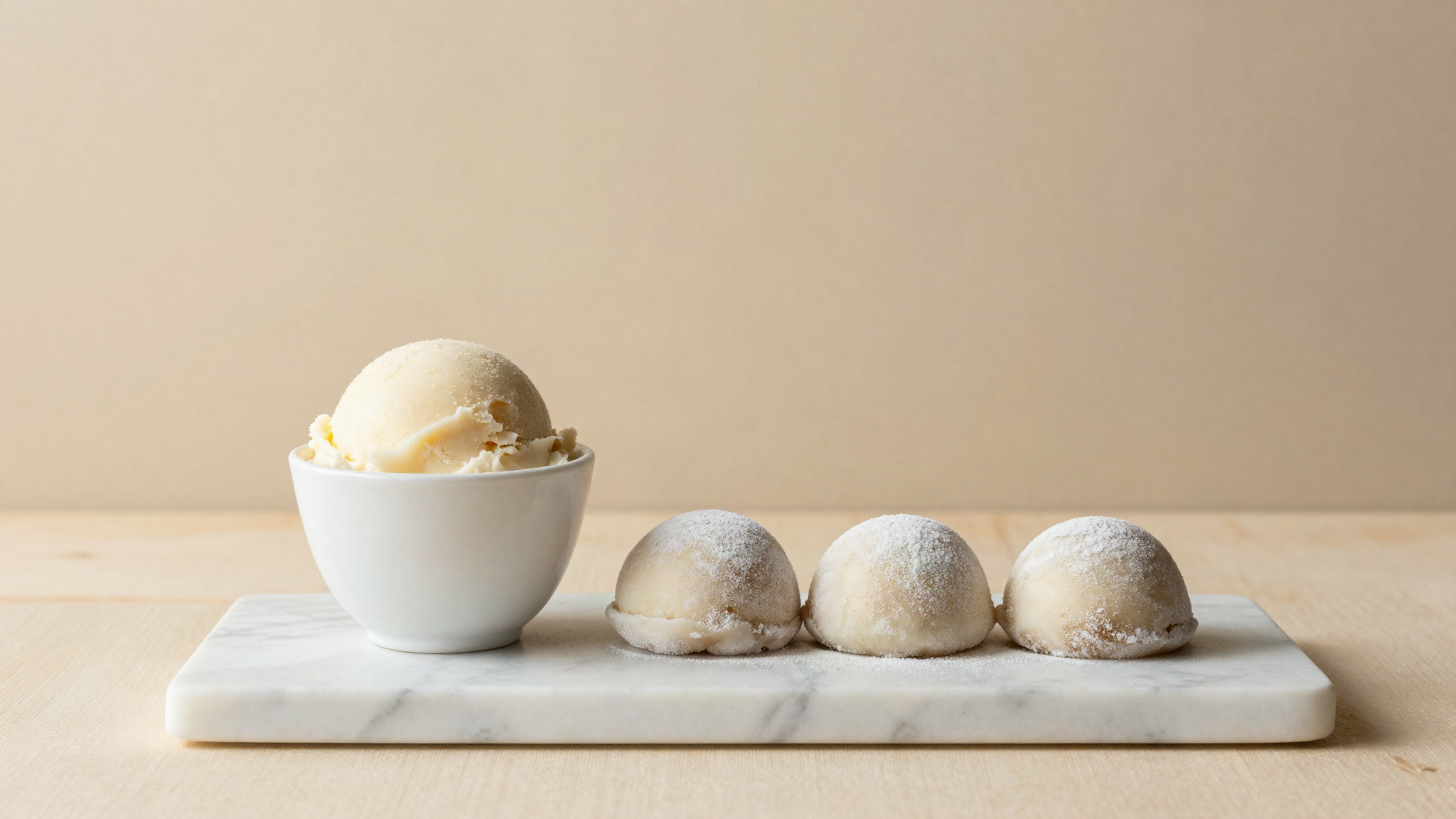 Cup of pale gelato beside three dusted mochi ice cream balls on a marble slab