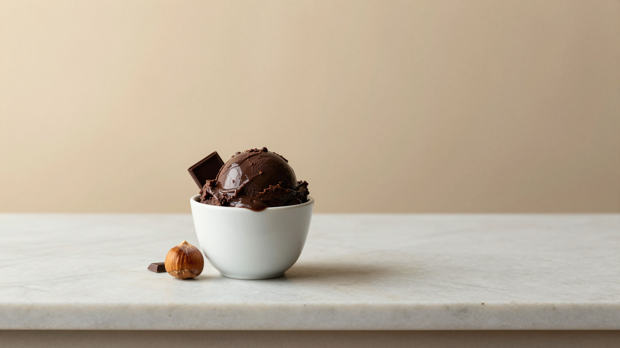 Small white ceramic cup with a scoop of dark glossy gianduia gelato beside a whole hazelnut and dark chocolate