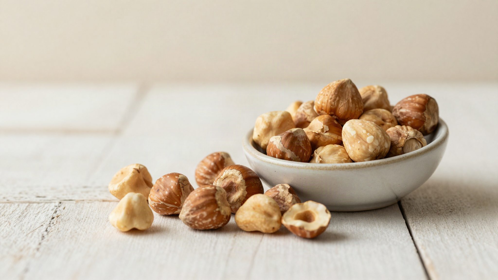 Whole toasted hazelnuts with papery skins spilling from a ceramic bowl onto aged wood