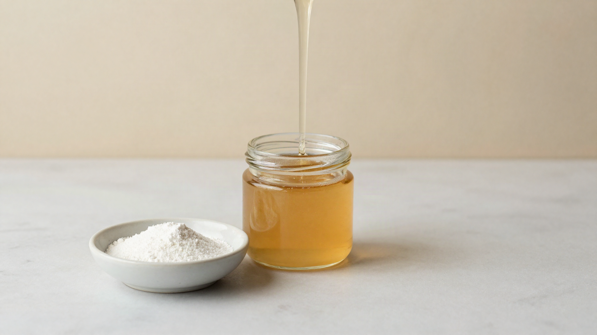 Glucose syrup as a hero ingredient on white marble, a small glass jar of thick clear syrup with a spoon drizzle beside a shallow dish of white glucose powder