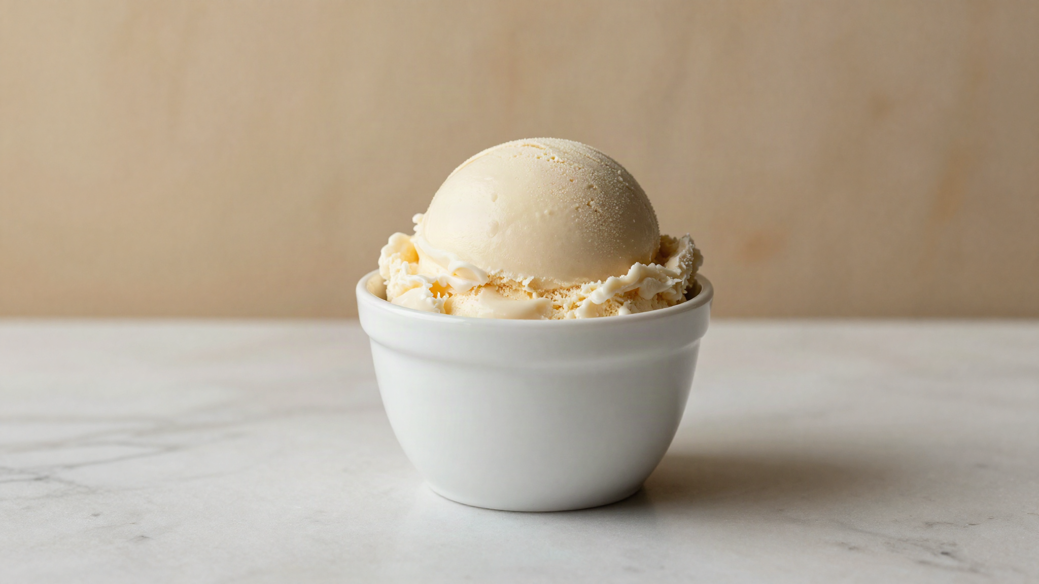 A single smooth scoop of pale ivory plant based gelato in a refined white ceramic cup on white marble