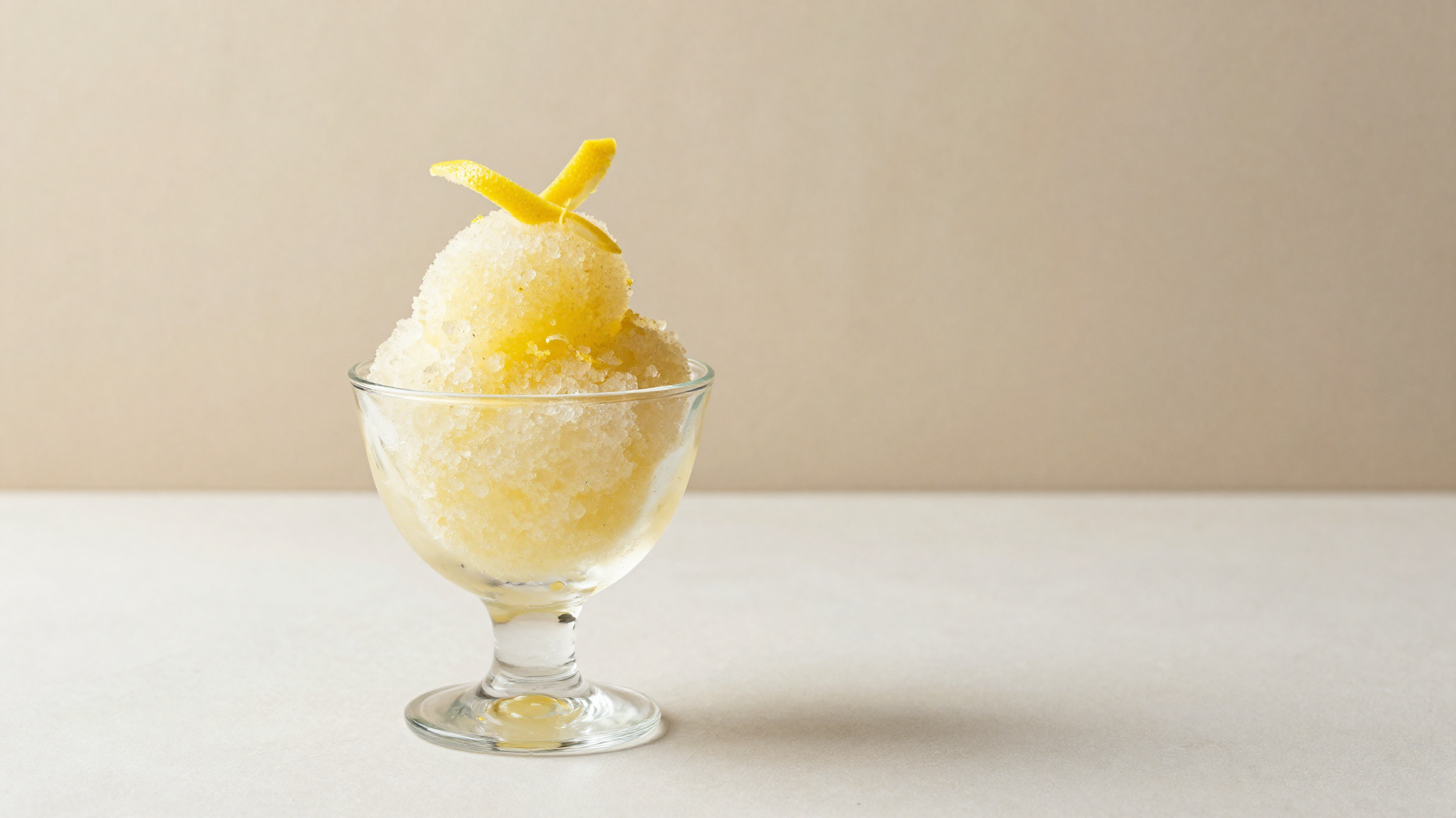 A chilled footed glass of Sicilian lemon granita with coarse pale-yellow ice crystals and a lemon twist
