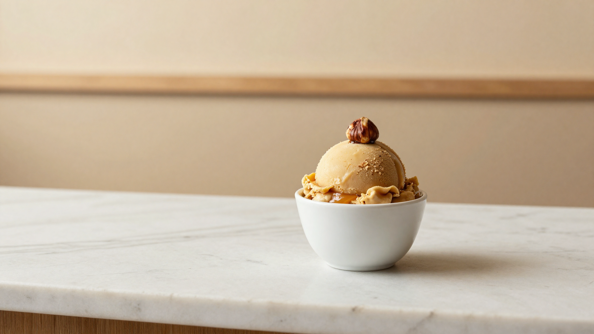 A scoop of warm caramel-brown hazelnut gelato in a white ceramic cup with a single roasted hazelnut garnish