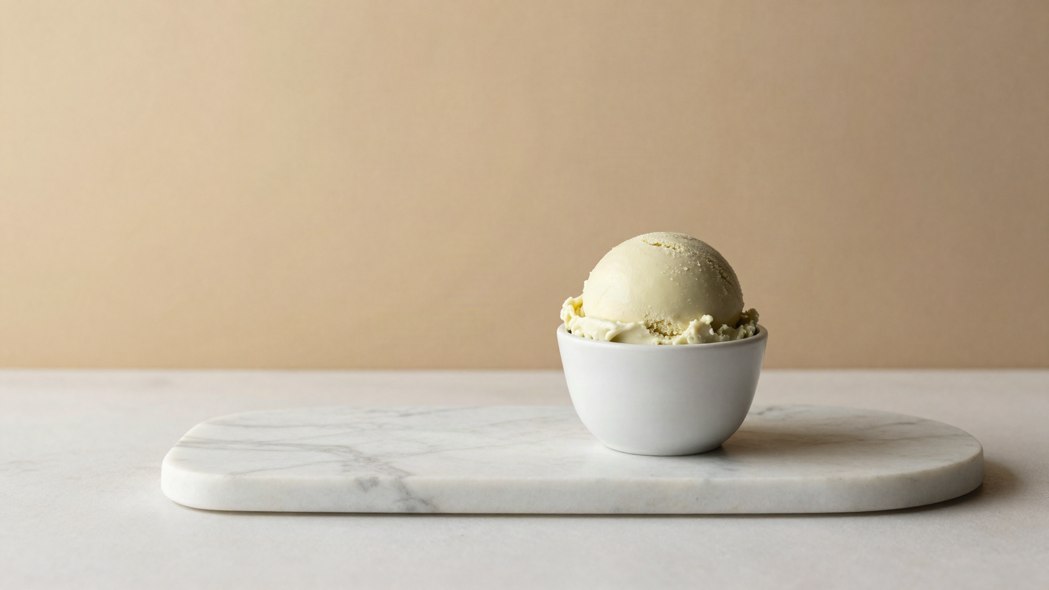 A single scoop of pale ivory green vegan gelato in a small white ceramic cup on marble