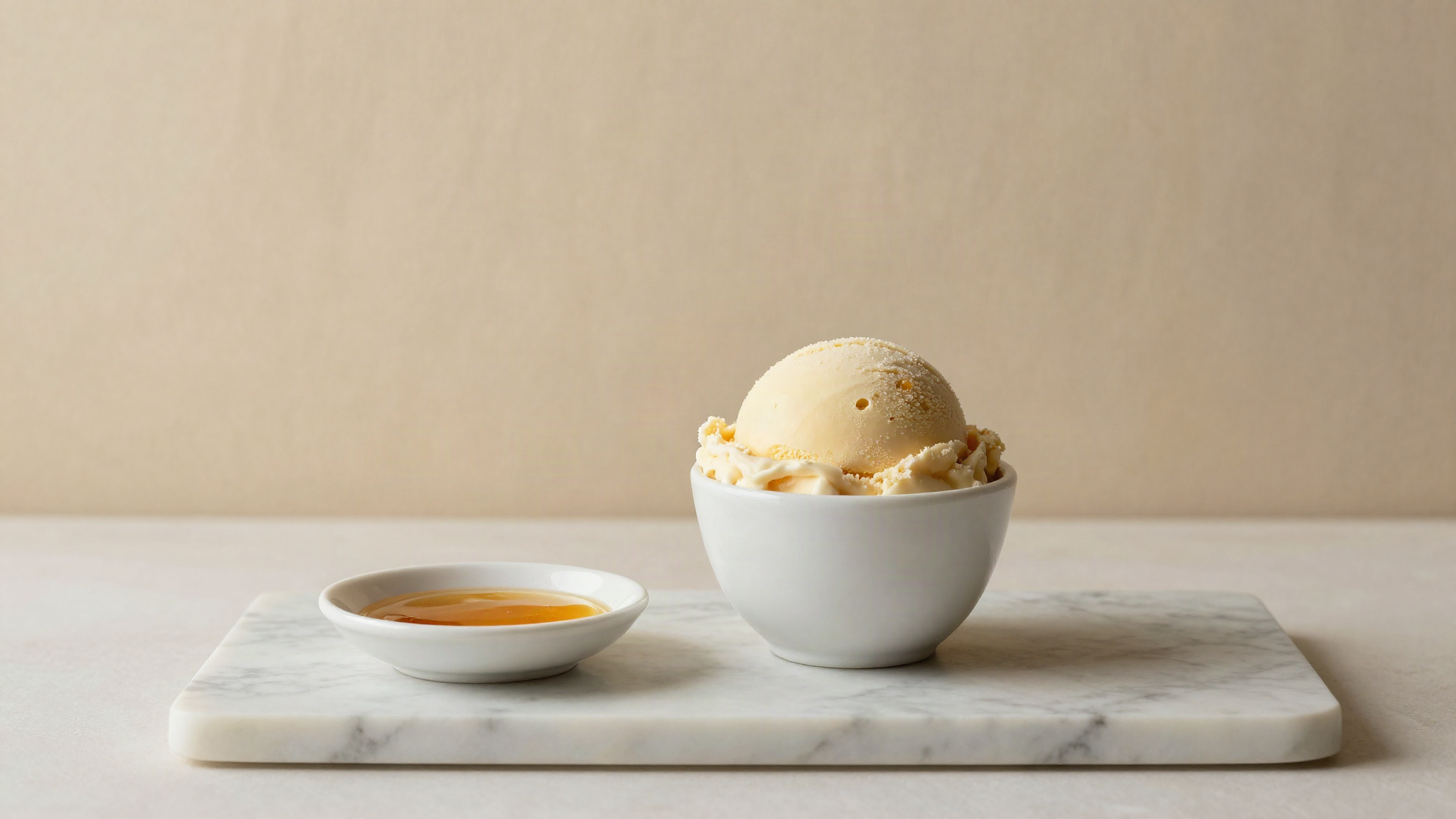A scoop of pale gelato in a small white ceramic cup on marble beside a shallow dish of amber honey catching the light