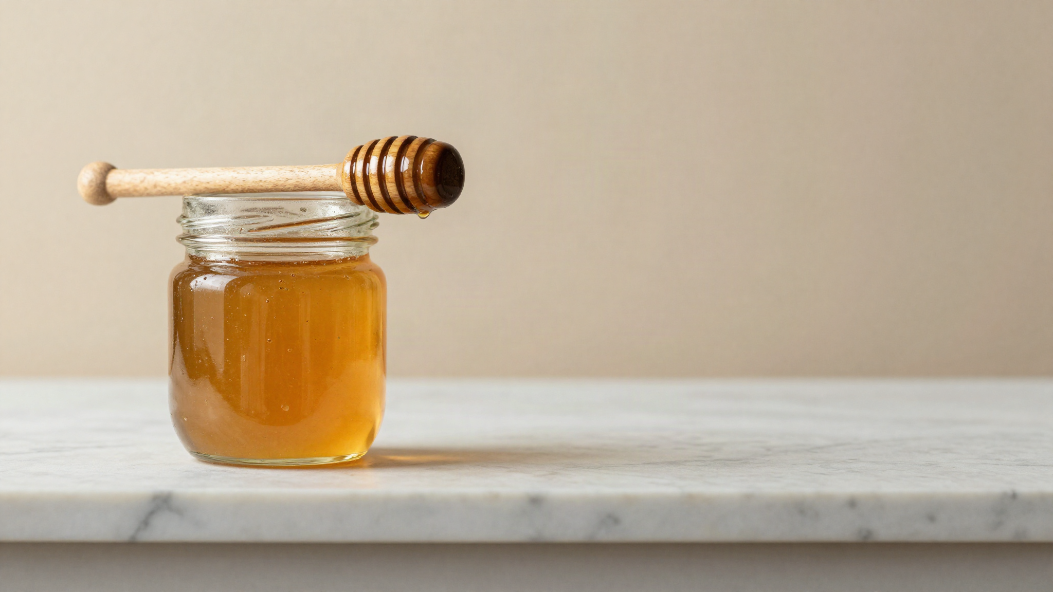 Raw amber honey in a clear glass jar with a wooden dipper resting across the rim on a marble counter