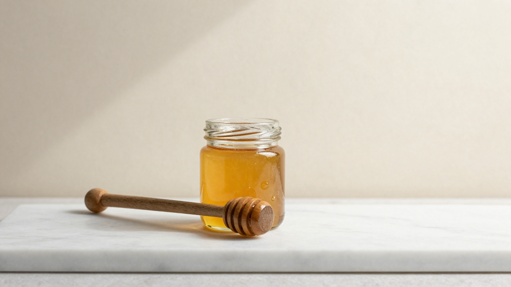A glass jar of golden honey with a wooden dipper on white marble, a hero ingredient for gelato
