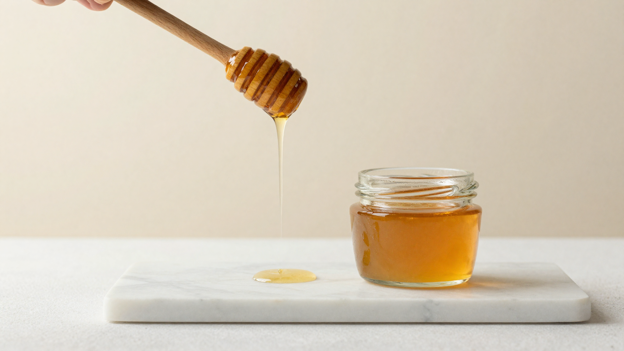 Golden honey drizzling from a wooden dipper beside a glass jar on marble