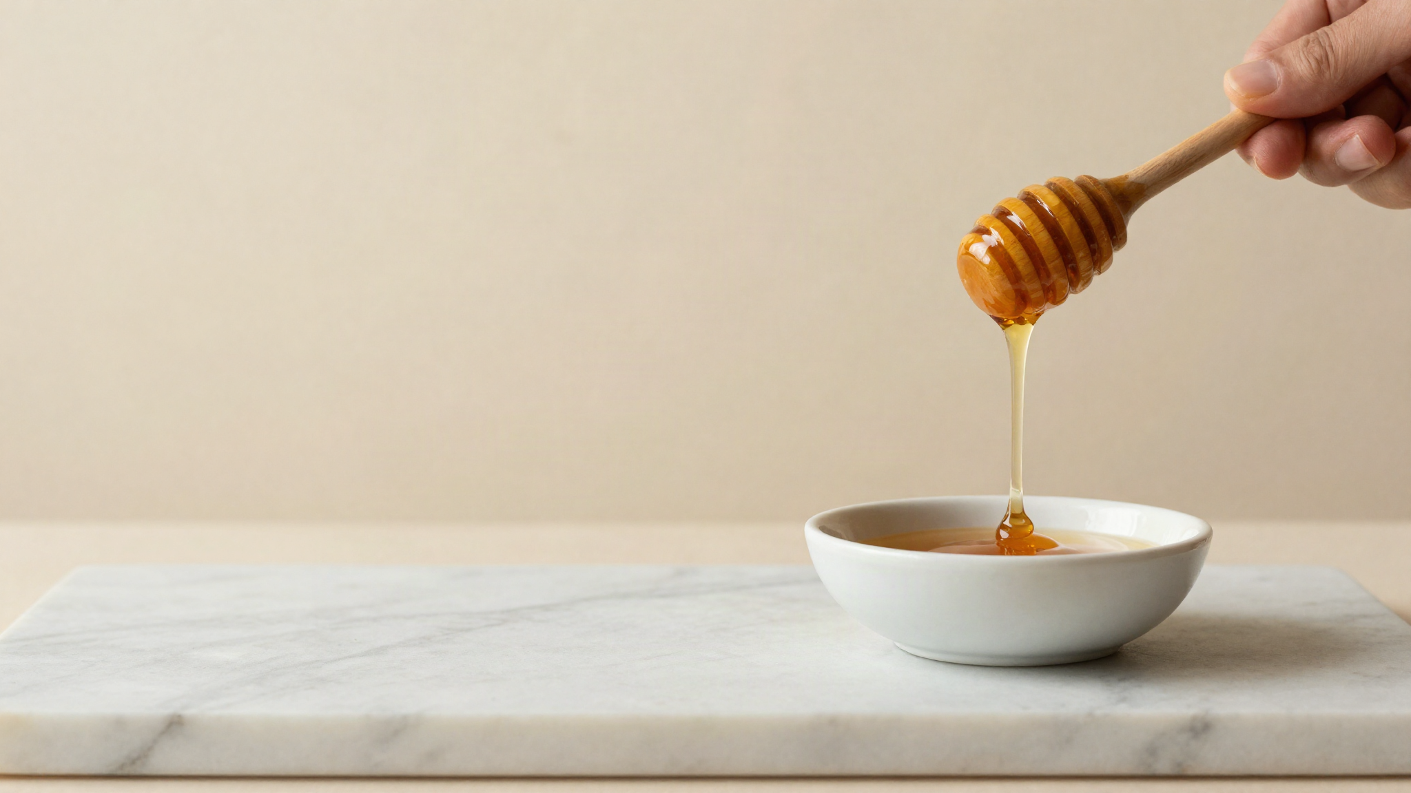 A honey dipper lifting a slow amber thread of honey over a small white ceramic bowl on marble