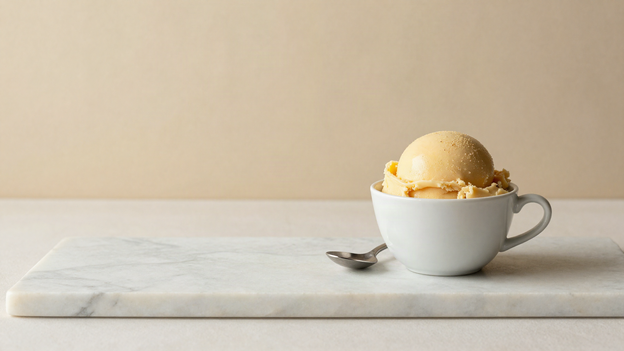 A small white ceramic cup of pale golden gelato with a spoon resting beside it on marble