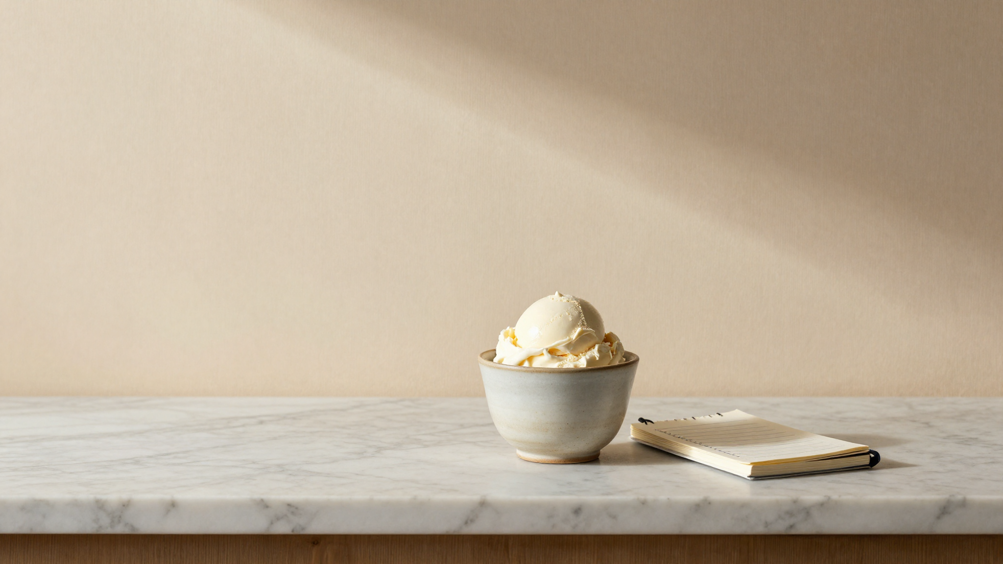 A refined ceramic cup holding a fresh small scoop of pale stracciatella gelato as a sample serving on a marble counter beside a small notebook with calculations