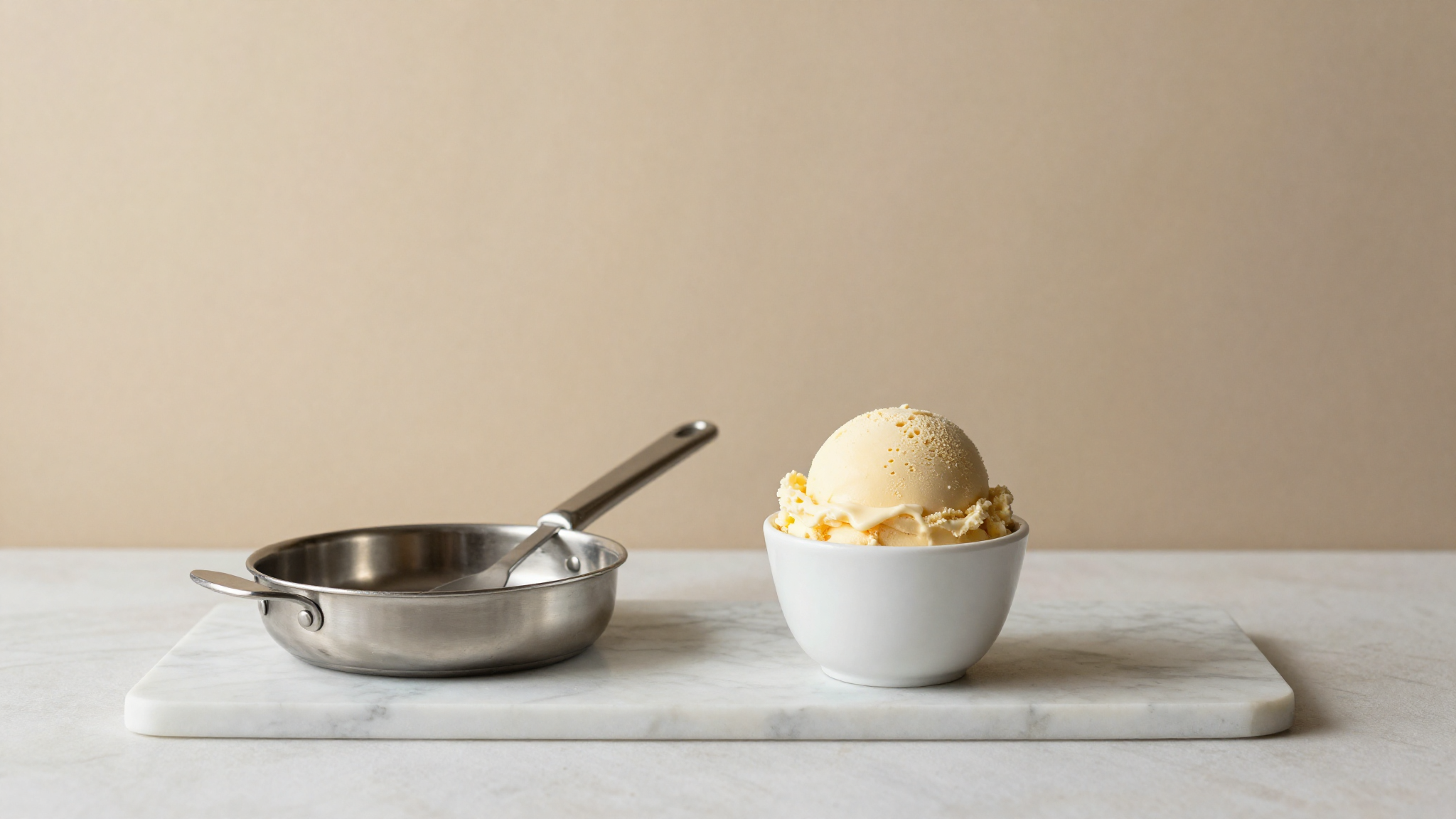 Freshly drawn scoop of dense Italian gelato in a white ceramic cup beside a curved stainless pan with a single paddle on marble