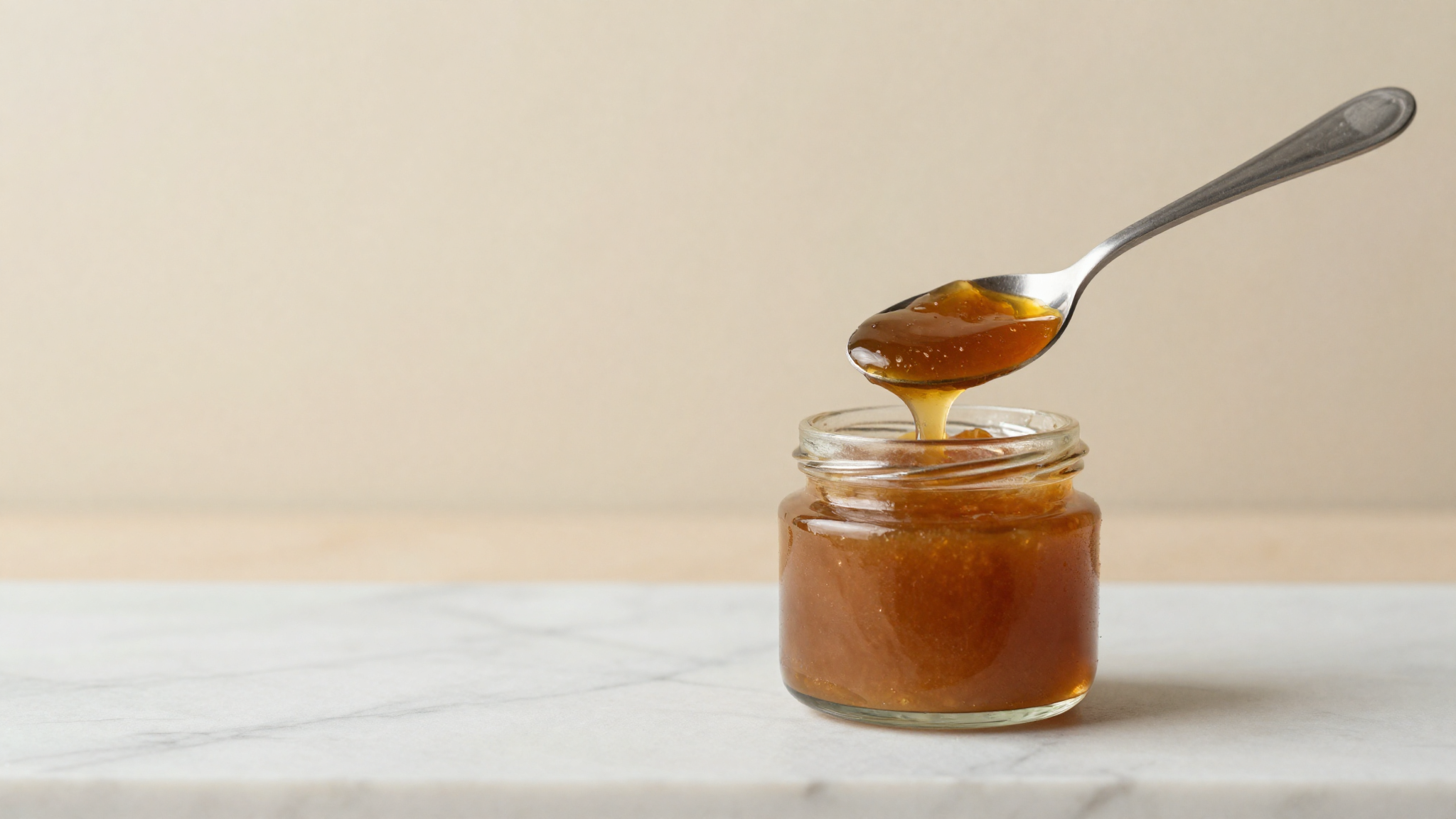 A spoon lifting thick amber invert sugar paste from a small glass jar on marble