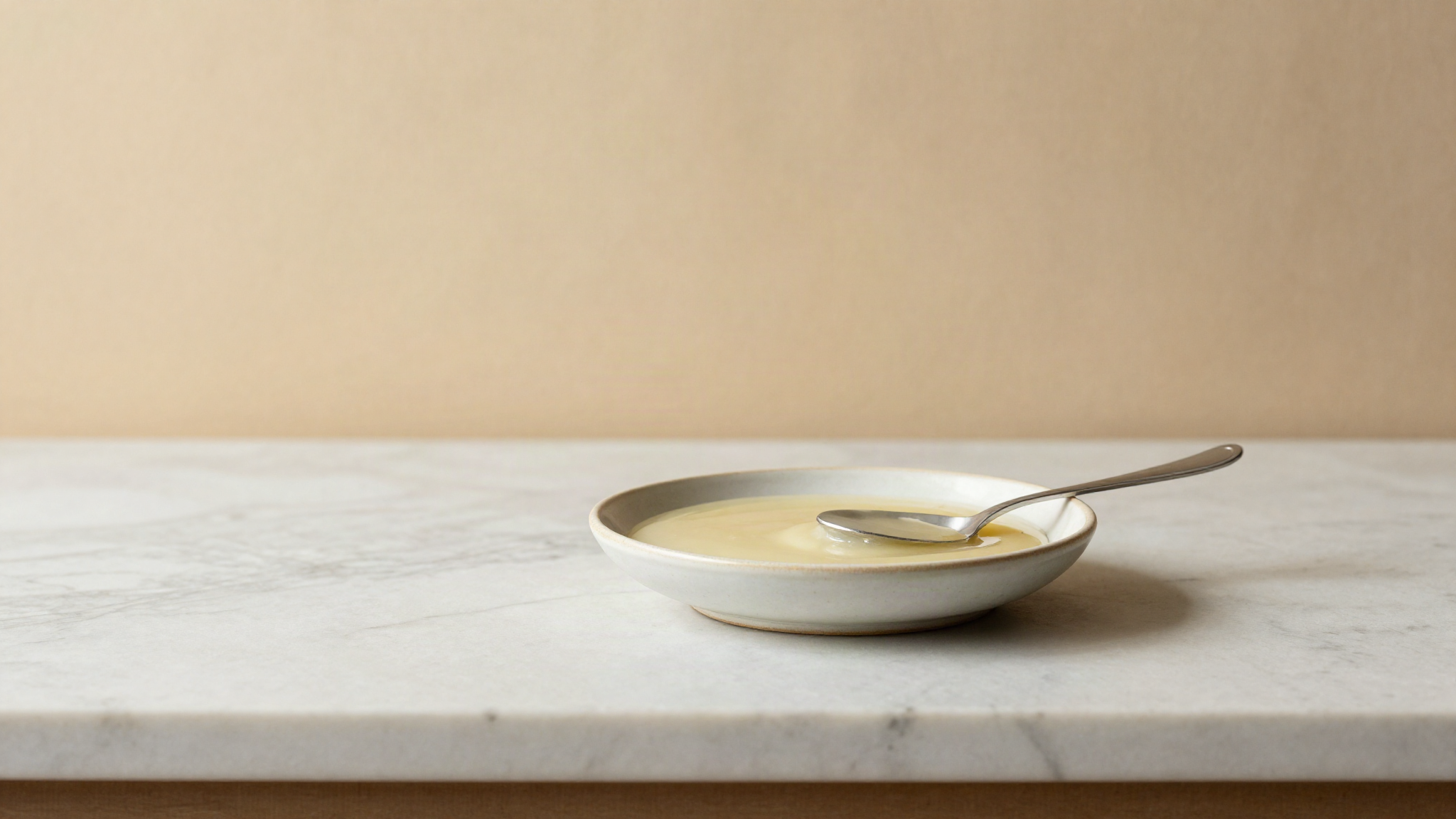 A shallow ceramic dish of thick pale invert sugar syrup catching light on a marble counter beside a small spoon