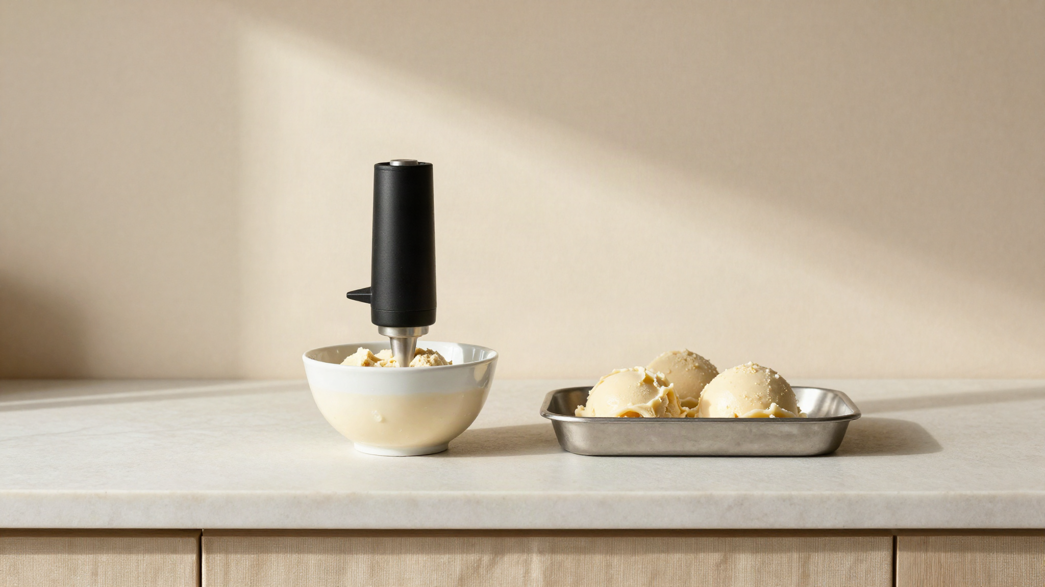 Small domestic food processor bowl beside a shallow metal tray of half-frozen pale gelato mix on cream linen