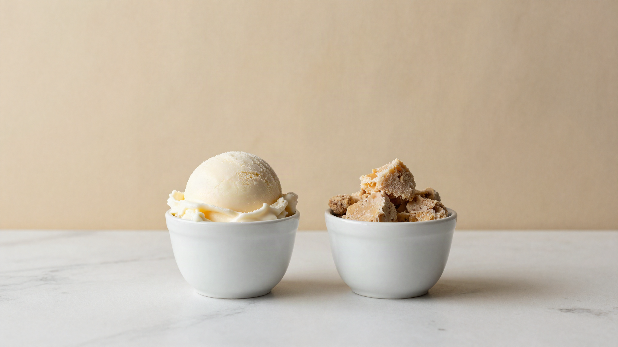 Two small white ceramic cups on marble, one with a dense smooth scoop of milk gelato, one with a coarse crumbly block
