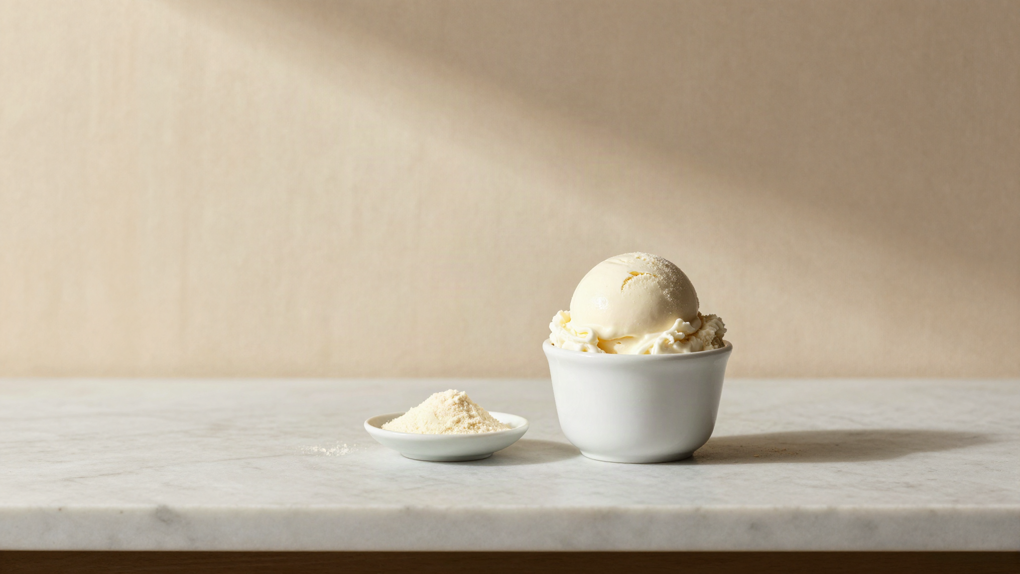 A single scoop of white fior di latte gelato in a small white ceramic cup on a marble counter, beside a small dish of pale stabilizer powder