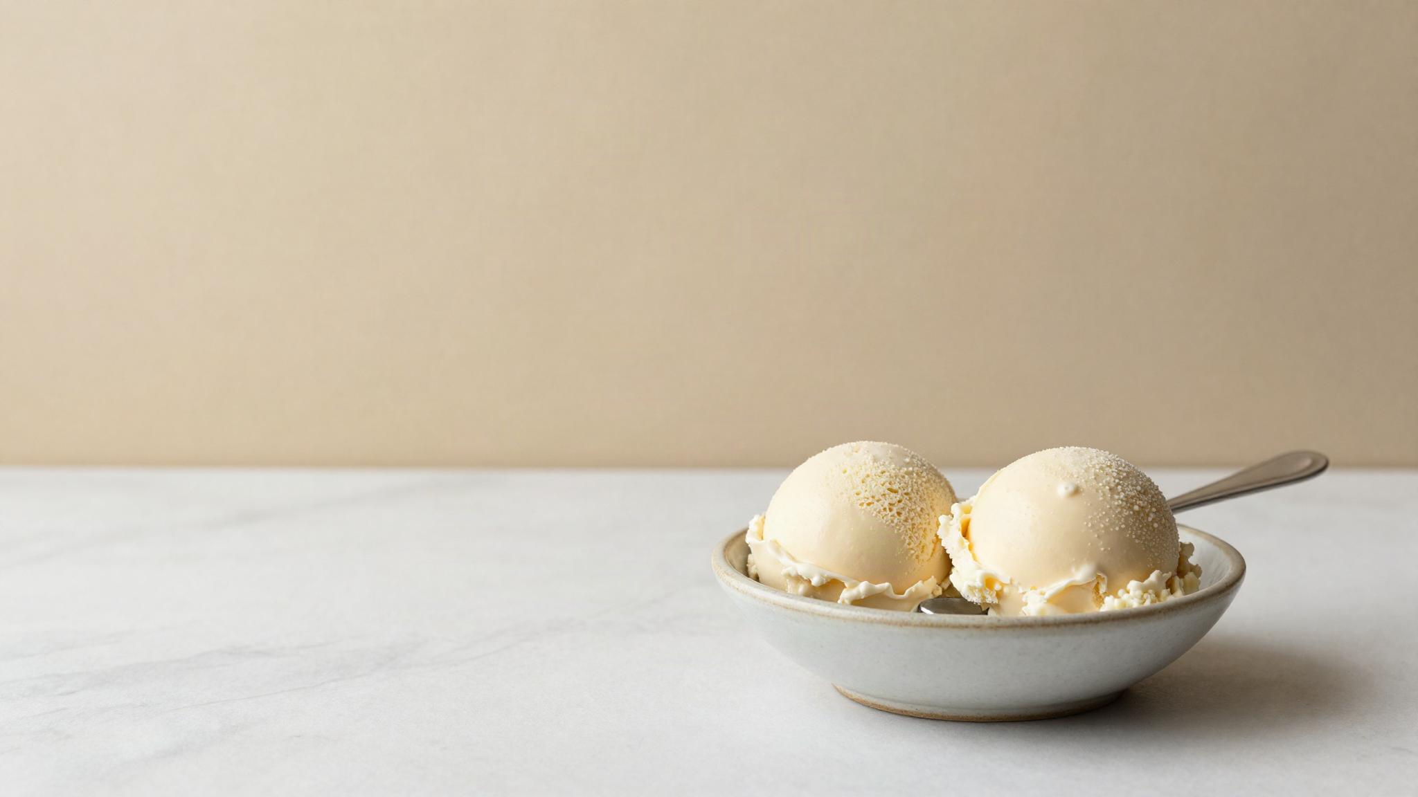 Two small scoops of pale ivory jasmine gelato in a chilled ceramic bowl with a small spoon on white marble
