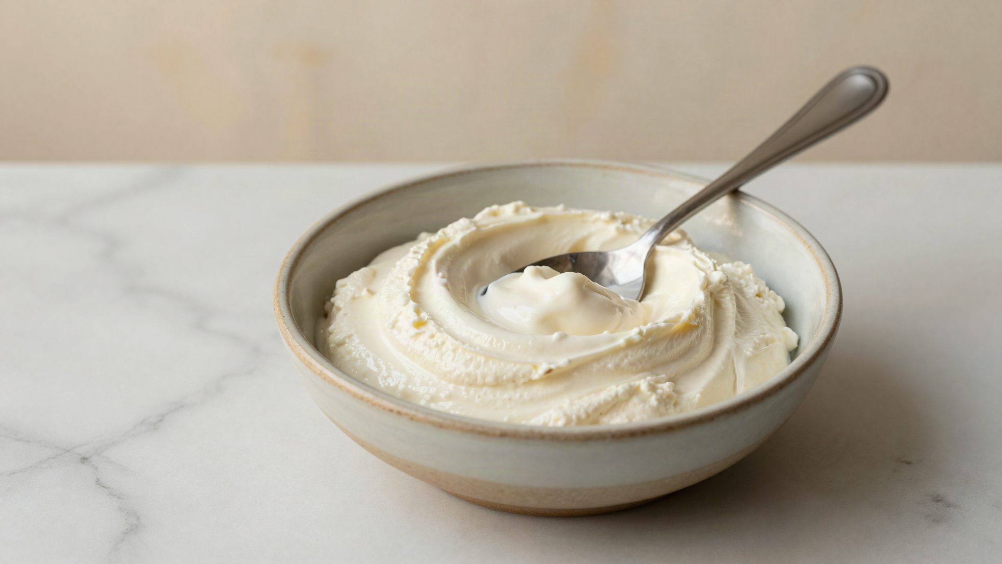 Bowl of thick strained labneh with a spoon swirl on a marble surface beside linen